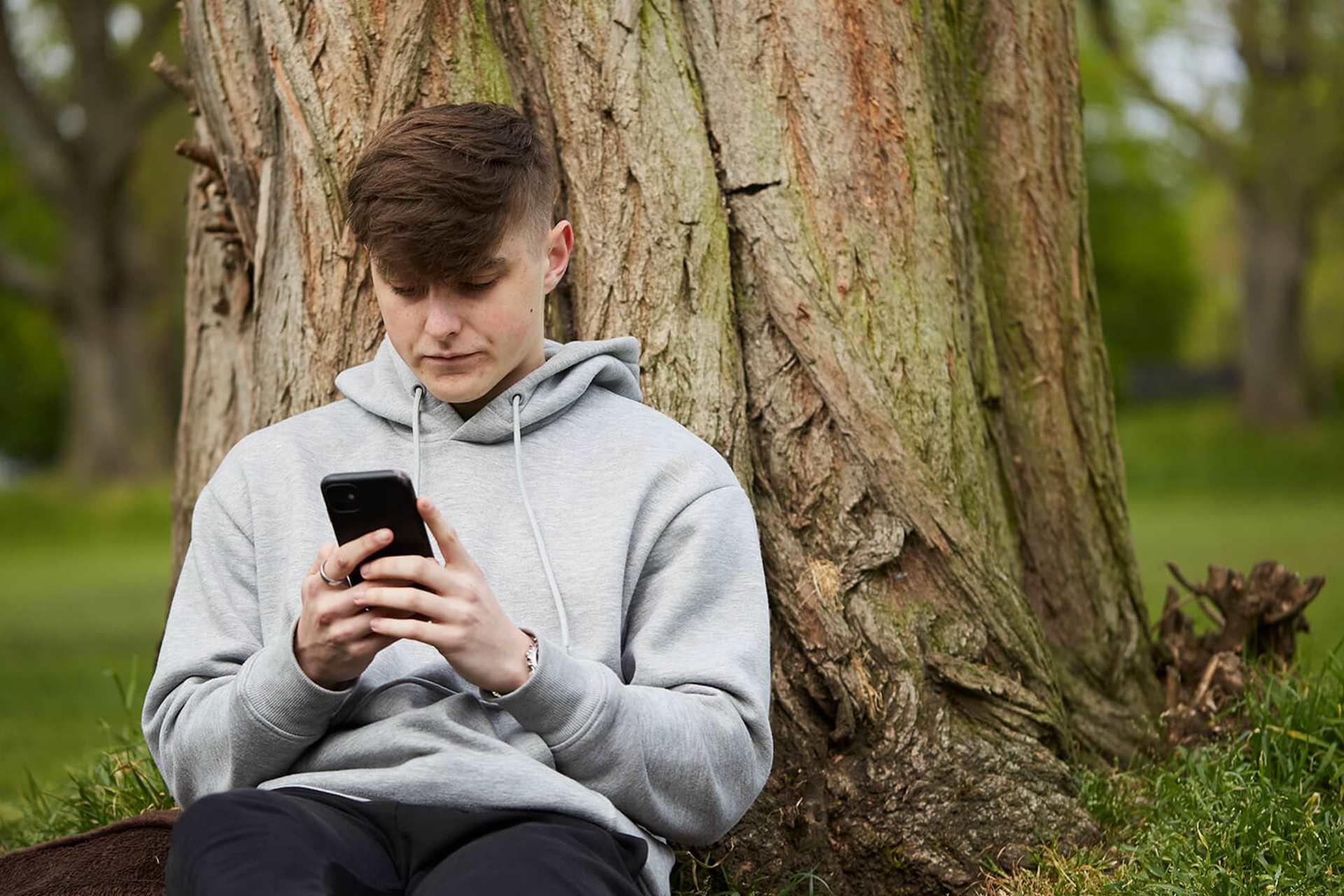 A young man wearing a grey hoodie and looking at his phone while he sits on the grass and leans against a tree.