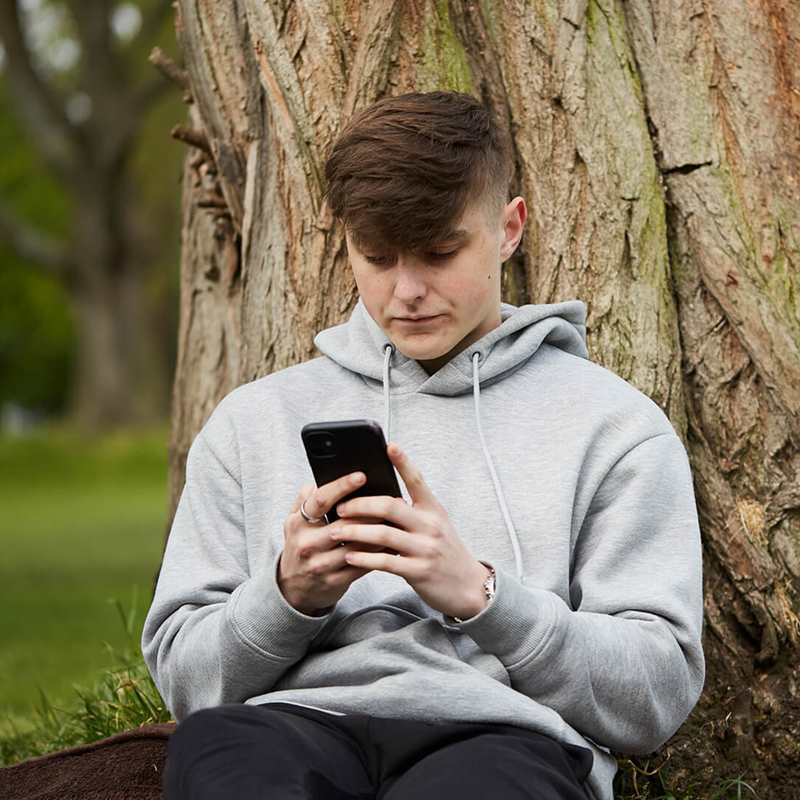 A young man wearing a grey hoodie and looking at his phone while he sits on the grass and leans against a tree.