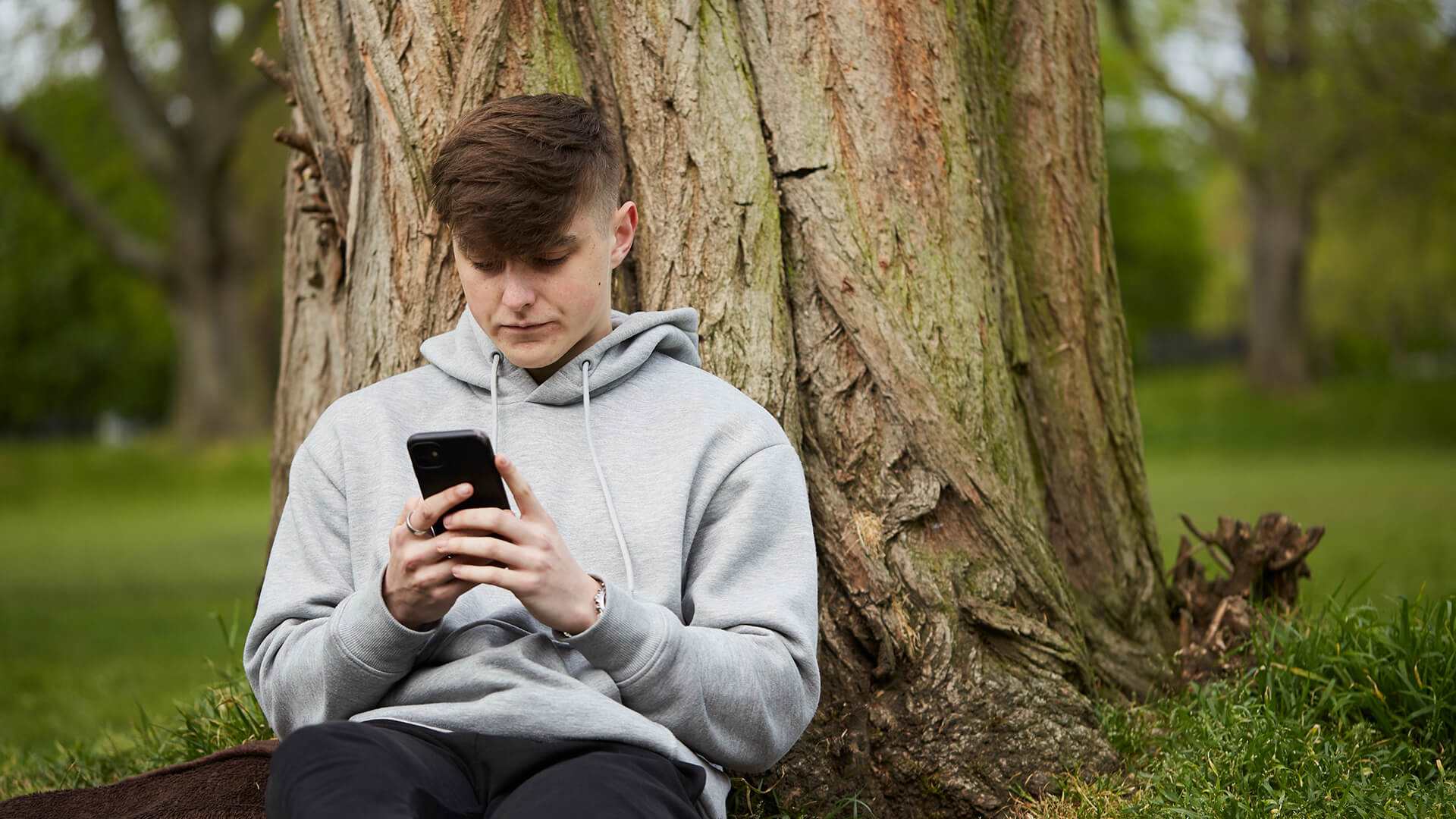 A young man wearing a grey hoodie and looking at his phone while he sits on the grass and leans against a tree.