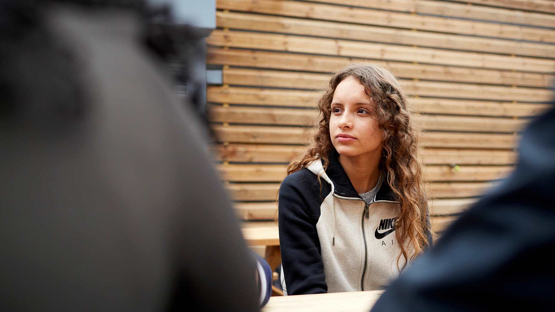 A girl with long, curly hair sits listening to her friends on a picnic bench.