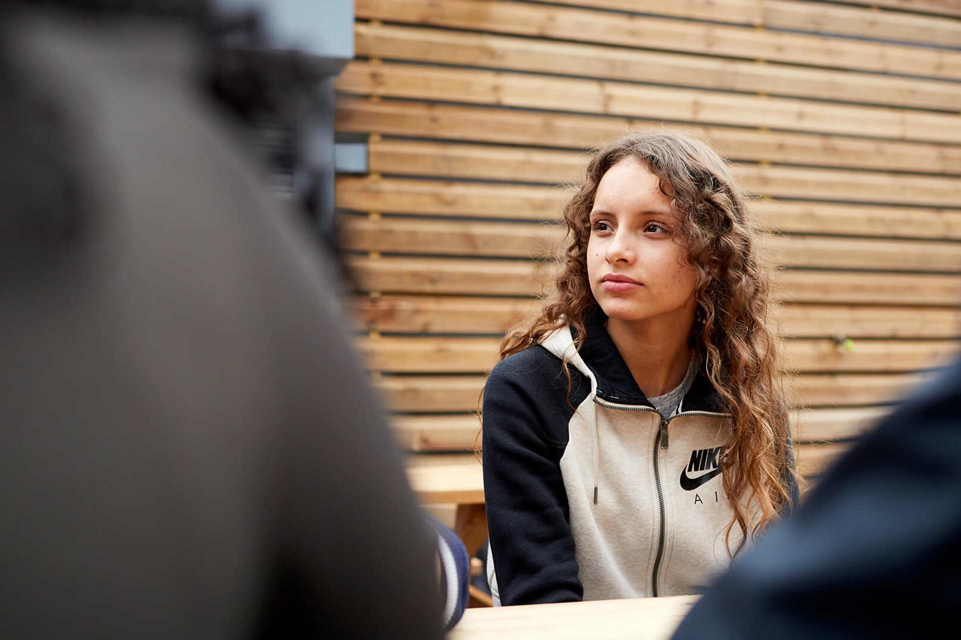 A girl with long, curly hair sits listening to her friends on a picnic bench.