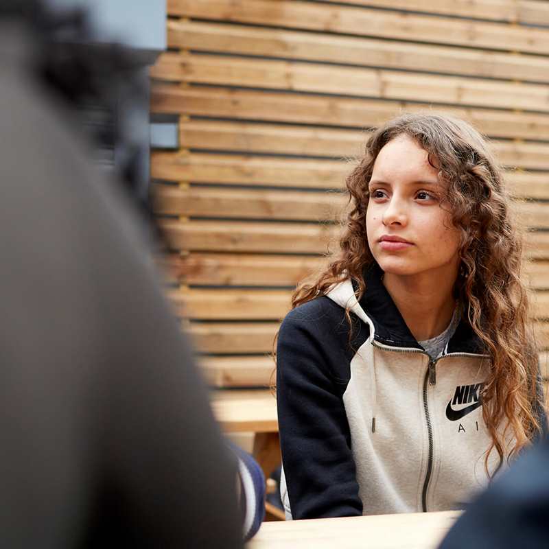 A girl with long, curly hair sits listening to her friends on a picnic bench.