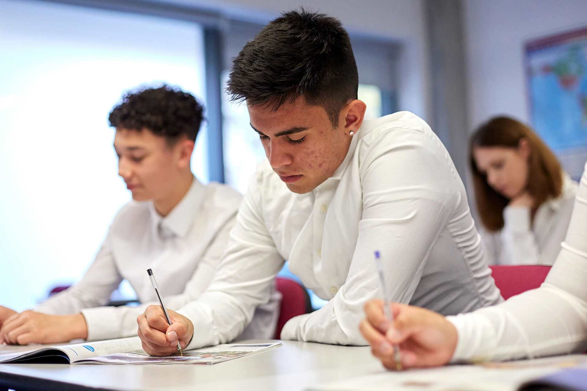 A group of students wearing school uniform sit at their desks in a classroom and write in their textbooks.