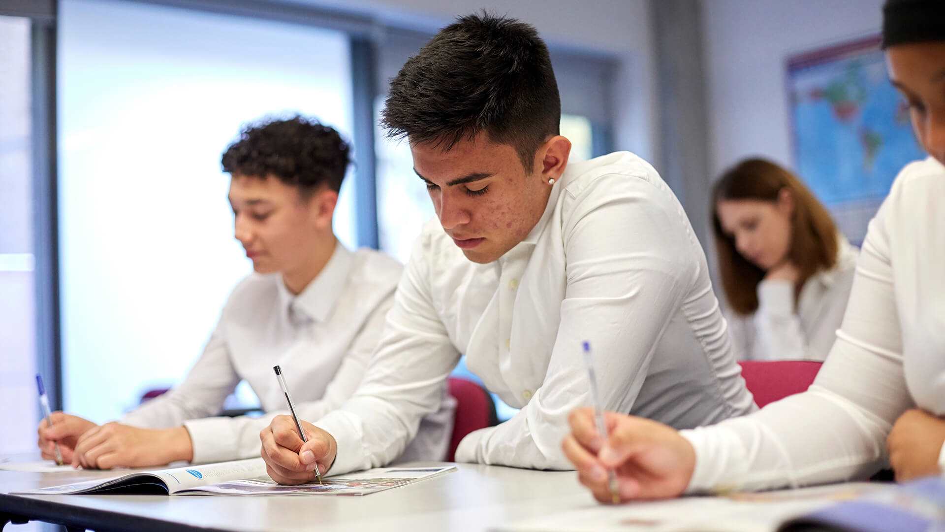 A group of students wearing school uniform sit at their desks in a classroom and write in their textbooks.