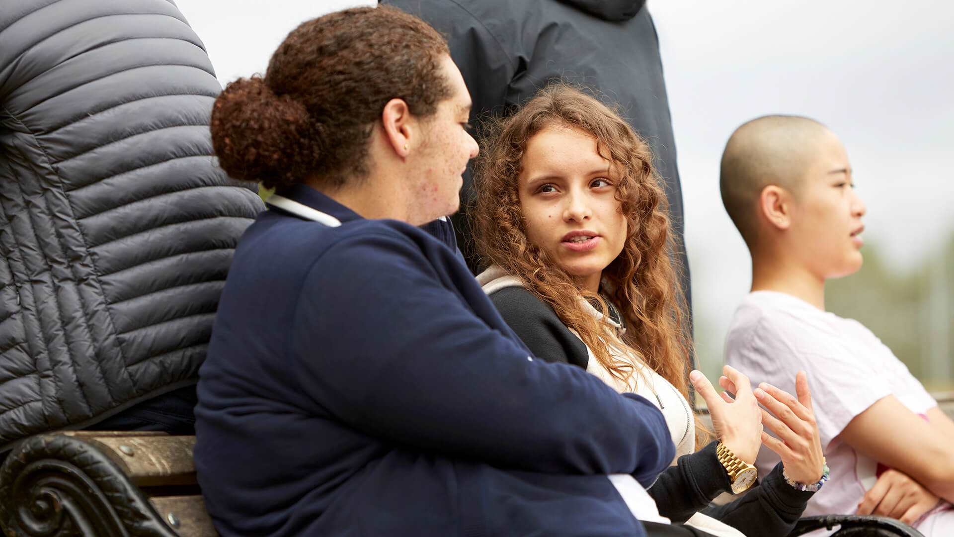 a-young-woman-with-a-curly-hair-talking-to-a-young-man-with-curly-hair-and-in-blue-jacket-while-sitting-on-a-bench-beside-another-girl-with-shaved-head-and-in-white-shirt-thinking-looking-serious-while-looking-away