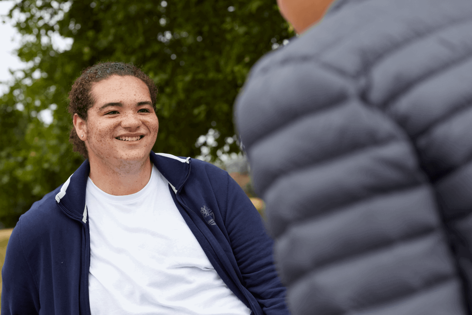 A young person smiling at their friend who is walking towards them.