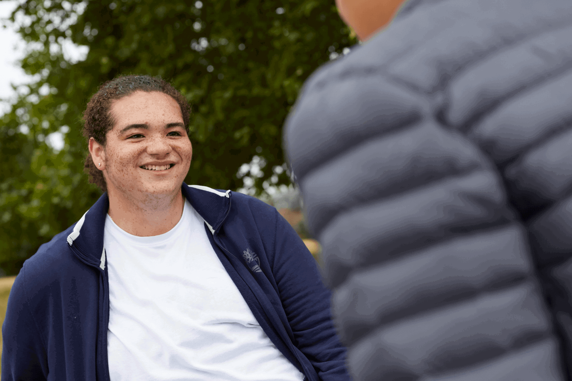 A young person smiling at their friend who is walking towards them.