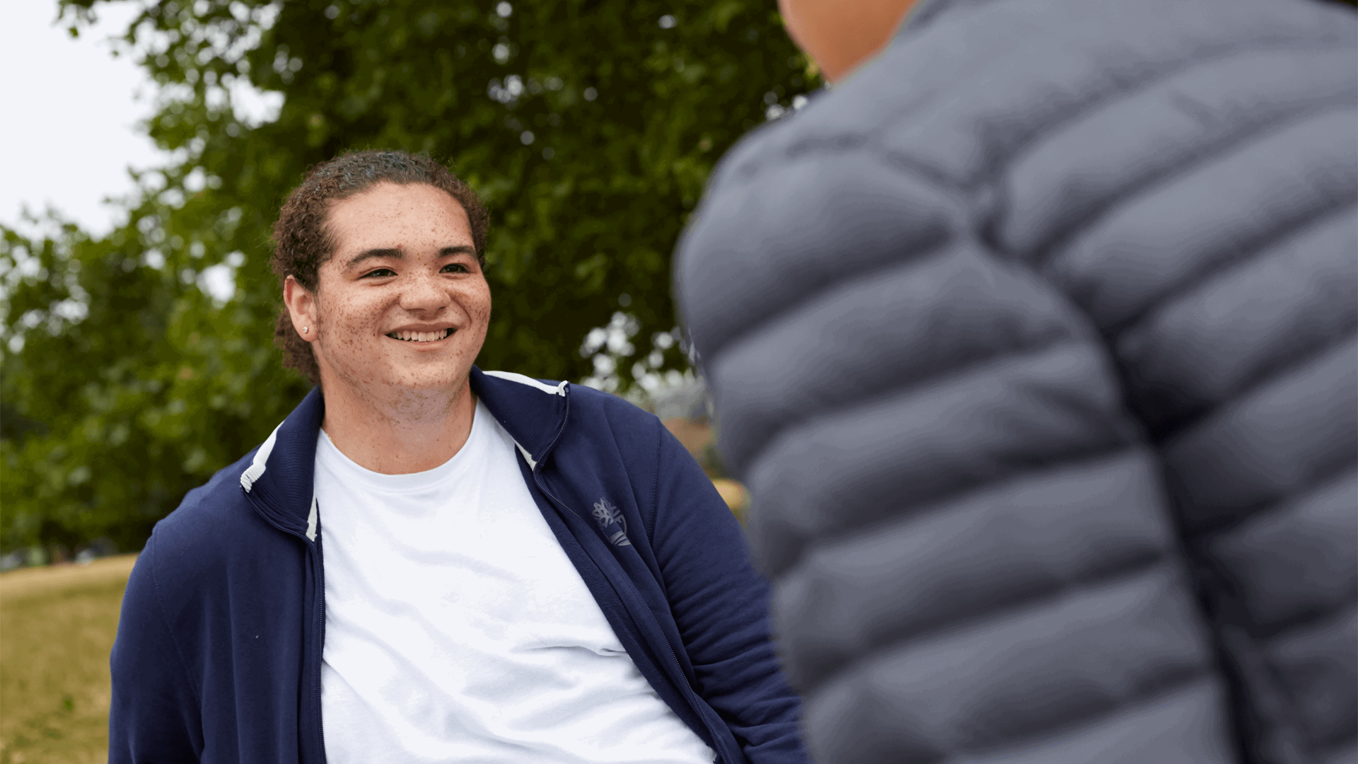 A young person smiling at their friend who is walking towards them.