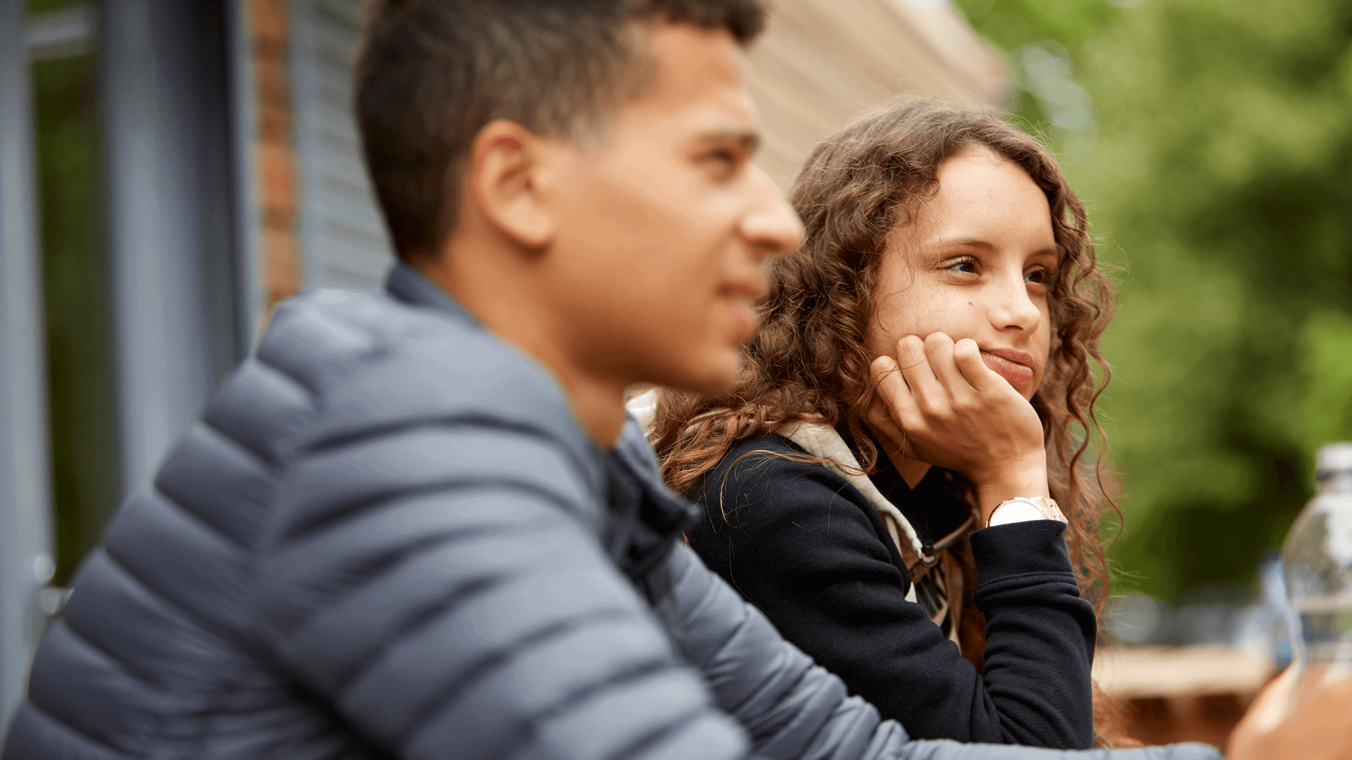 A girl with curly hair sits with her hand on her chin thinking, while a boy sits beside her wearing a grey jacket.