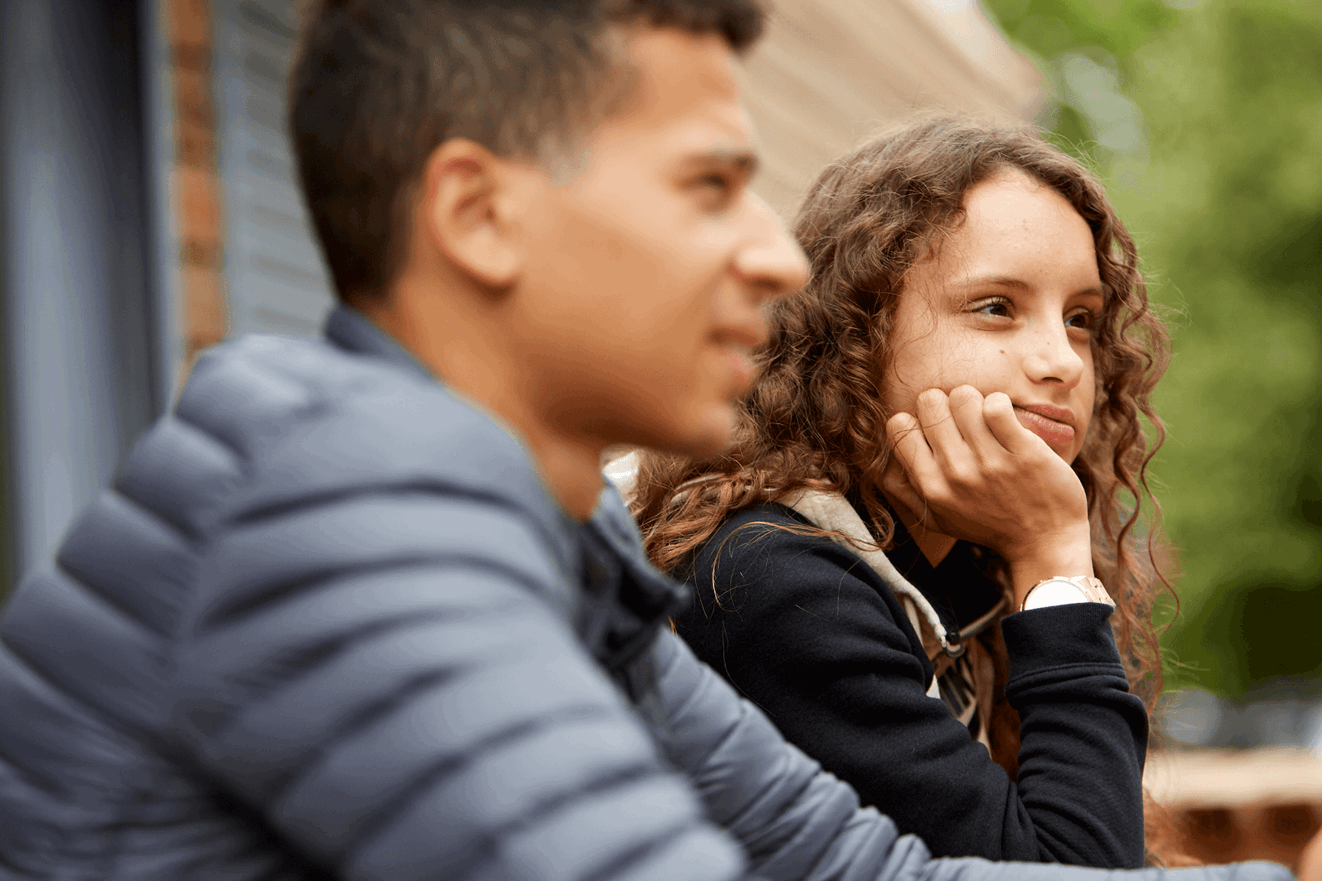 A girl with curly hair sits with her hand on her chin thinking, while a boy sits beside her wearing a grey jacket.