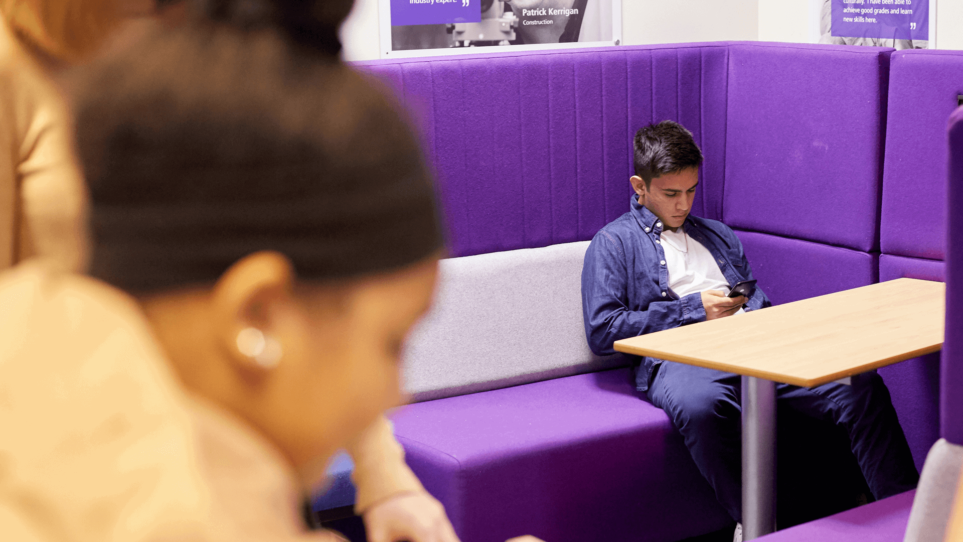 wide shot of a boy looking at his phone while sitting on a chair and table in the corner of the room while friends on foreground are having fun