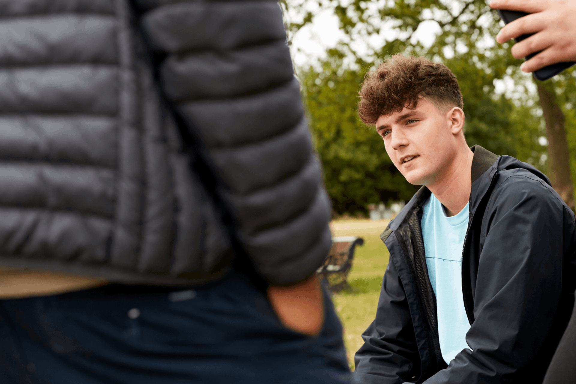 A young person sitting on a bench in a park with friends standing around him.