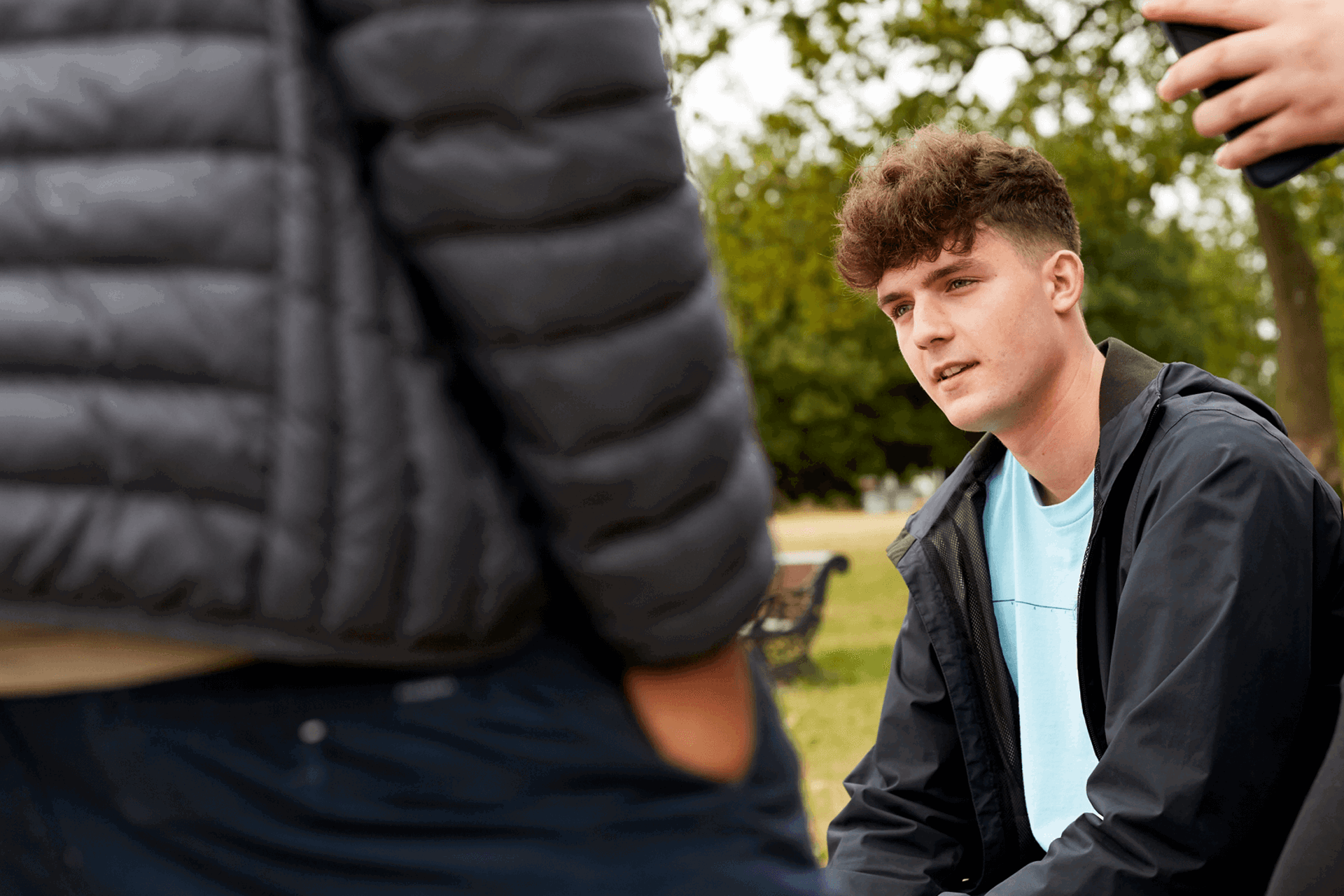 A young person sitting on a bench in a park with friends standing around him.