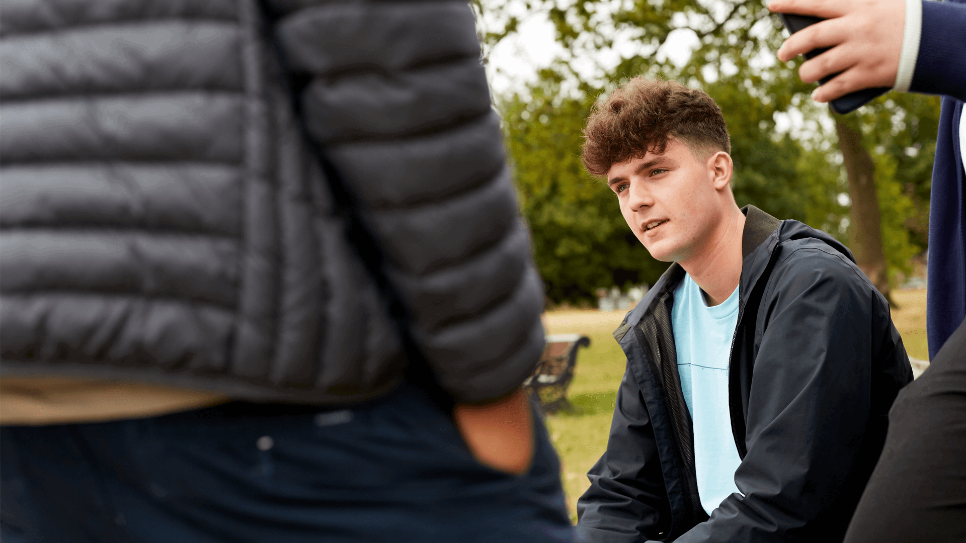 A young person sitting on a bench in a park with friends standing around him.