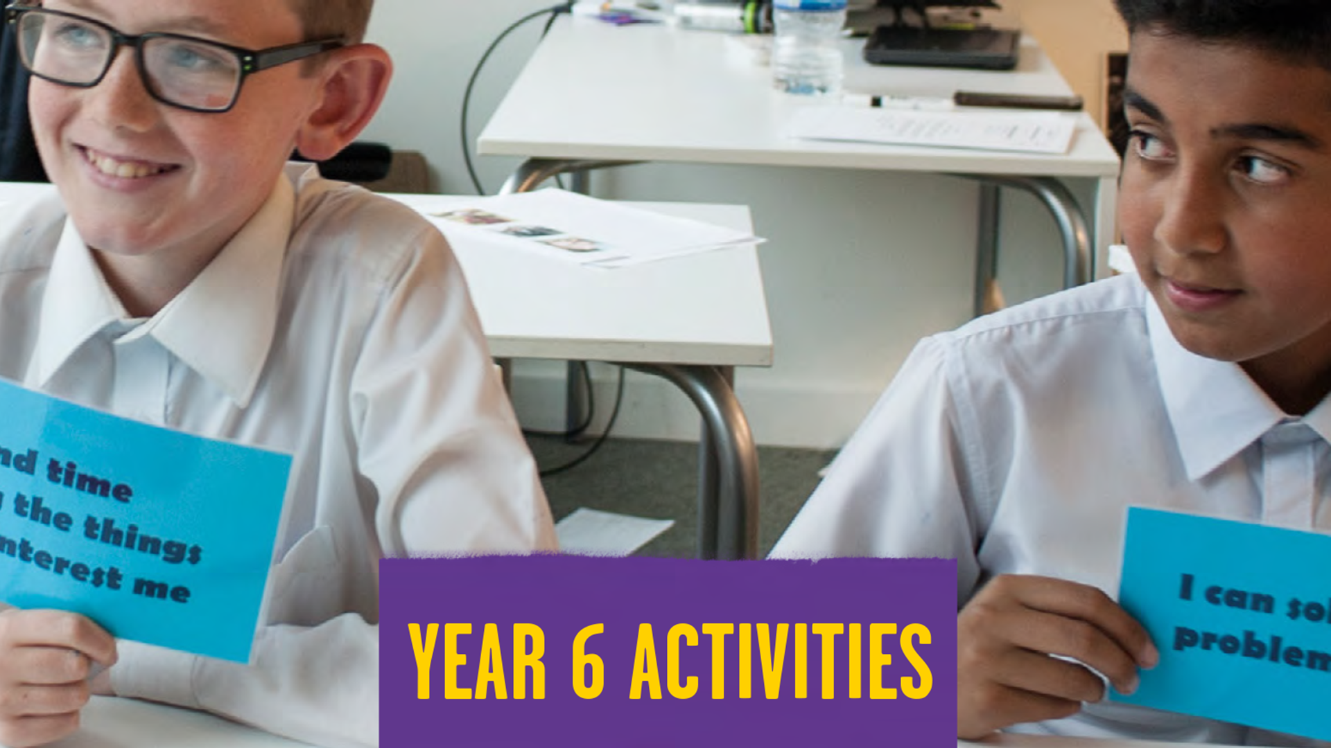 Two students sit next to each at a desk in a classroom holding blue cards with different statements on. In the middle of the students, there's a purple square with yellow writing. It says 'year 6 activities'.