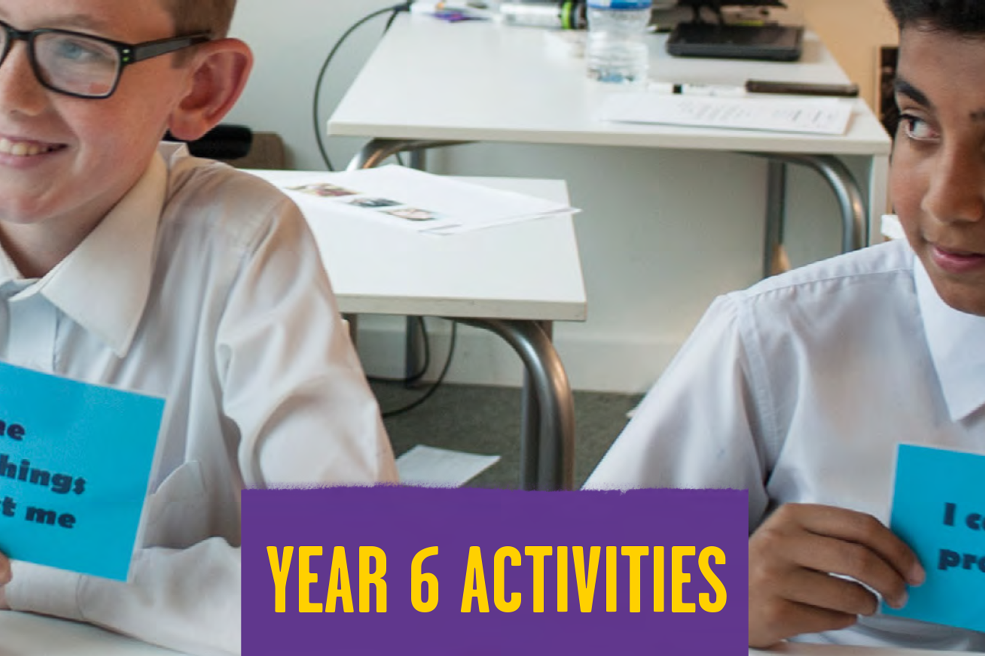Two students sit next to each at a desk in a classroom holding blue cards with different statements on. In the middle of the students, there's a purple square with yellow writing. It says 'year 6 activities'.
