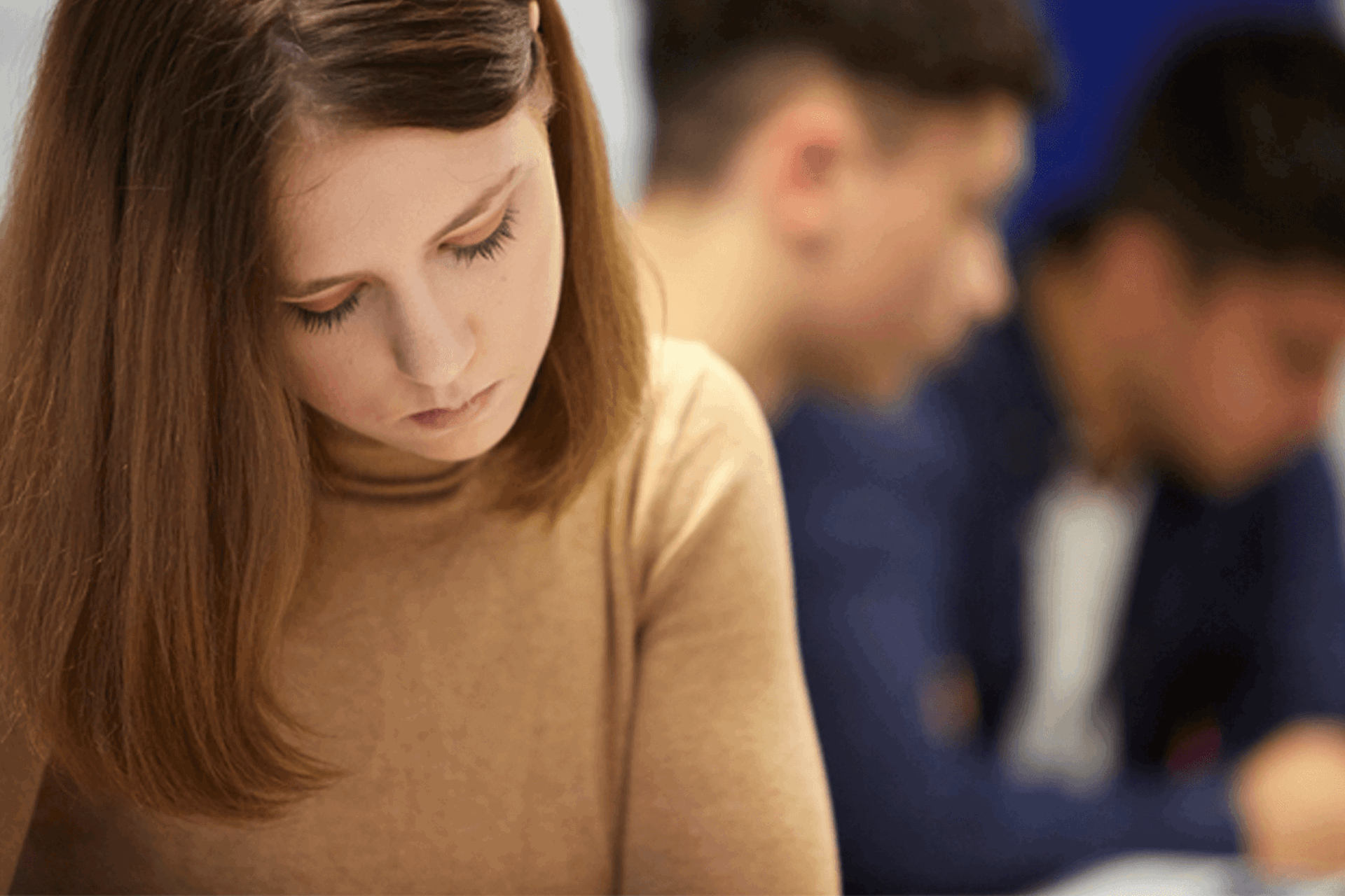 a young woman in brown jumper looking down with sad expression while two young men are blurred on the background