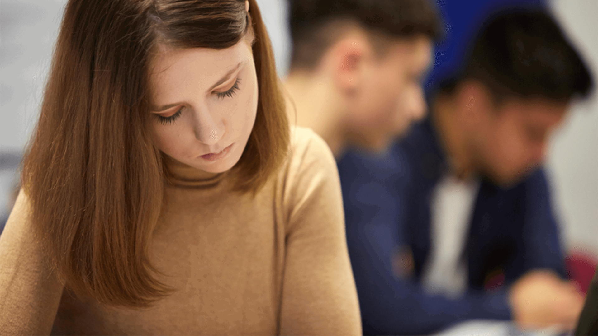 a young woman in brown jumper looking down with sad expression while two young men are blurred on the background