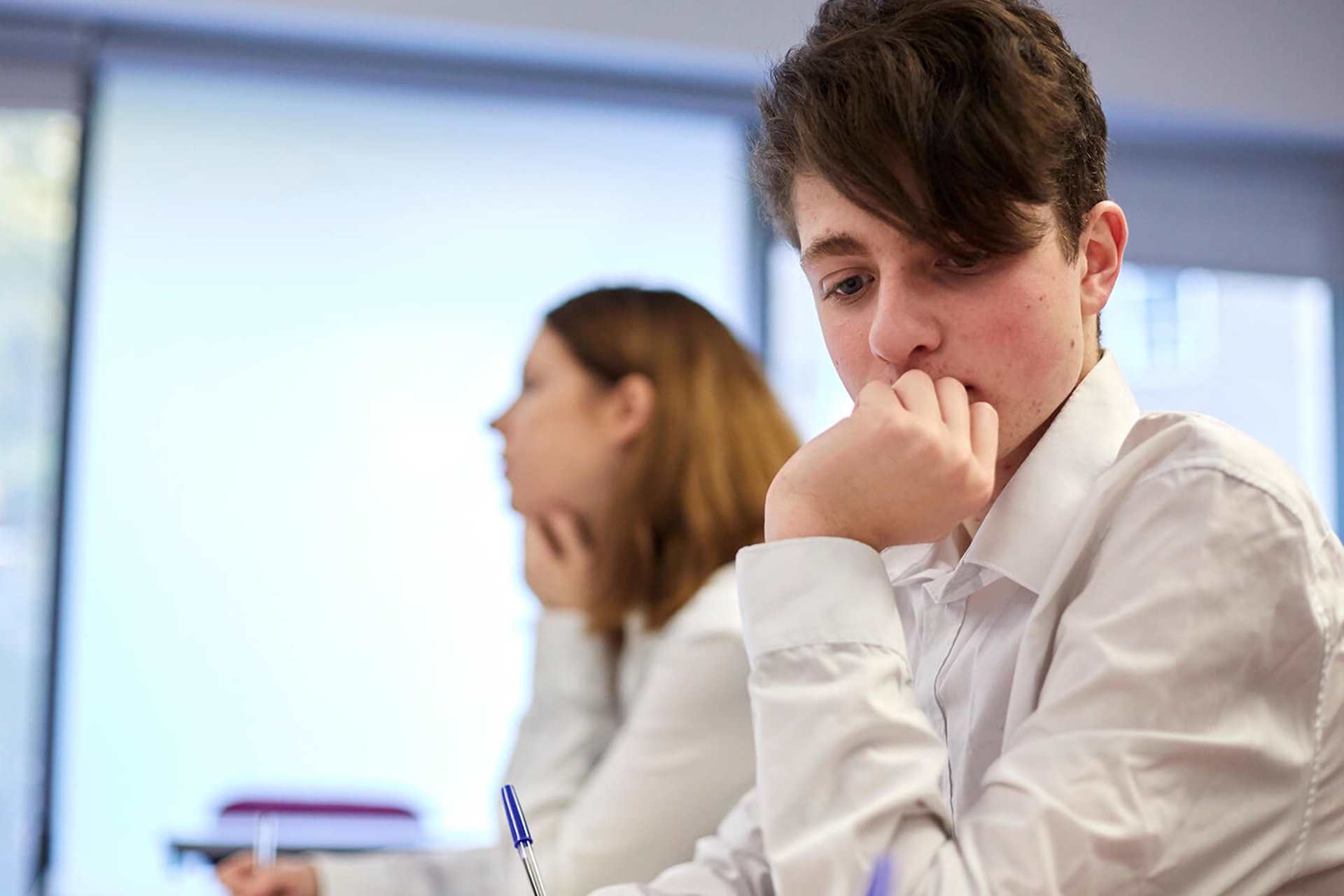 A student wearing uniform sits on a desk lost in a thought with their hand over their mouth, they sit next to another student who is focused on the lesson.