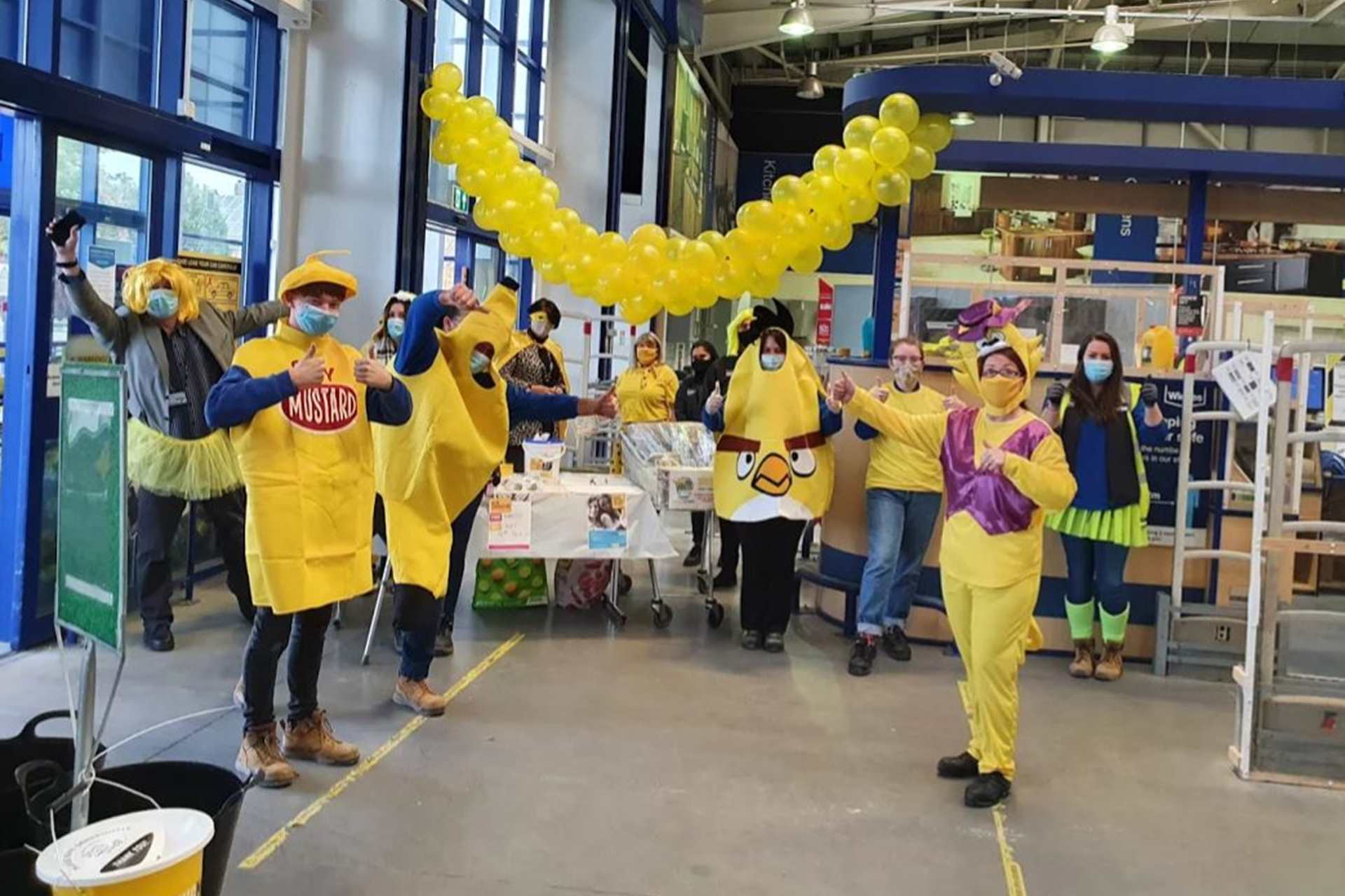 Members of staff from the Wickes branch in Newcastle are dressed up in yellow clothes and fancy dress outfits including a banana, a yellow bird, and mustard. They all standing round a table with their thumbs up facing the camera.