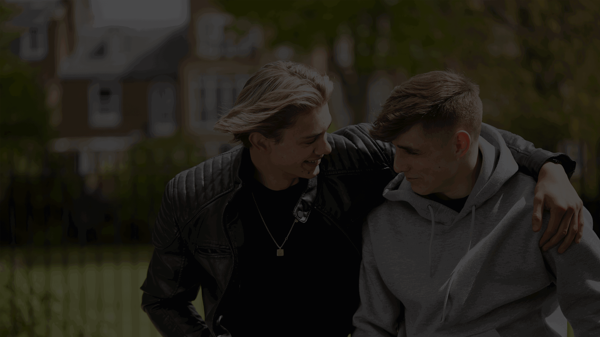 Two young people are talking and looking at each other. The young person on the left has got their arm around the other young person while smiling at them. In the background is a park with houses behind the park.