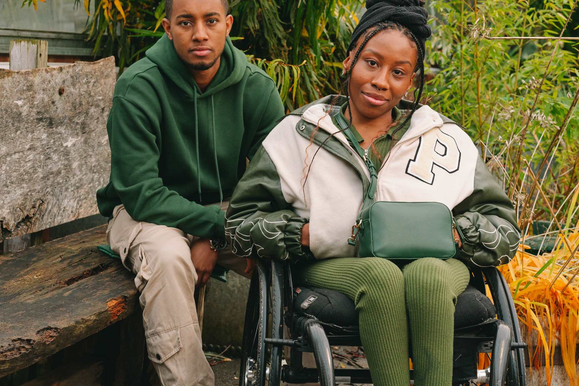 A young Black woman in a wheelchair and a young Black man on a bench, both staring at the camera looking serious.