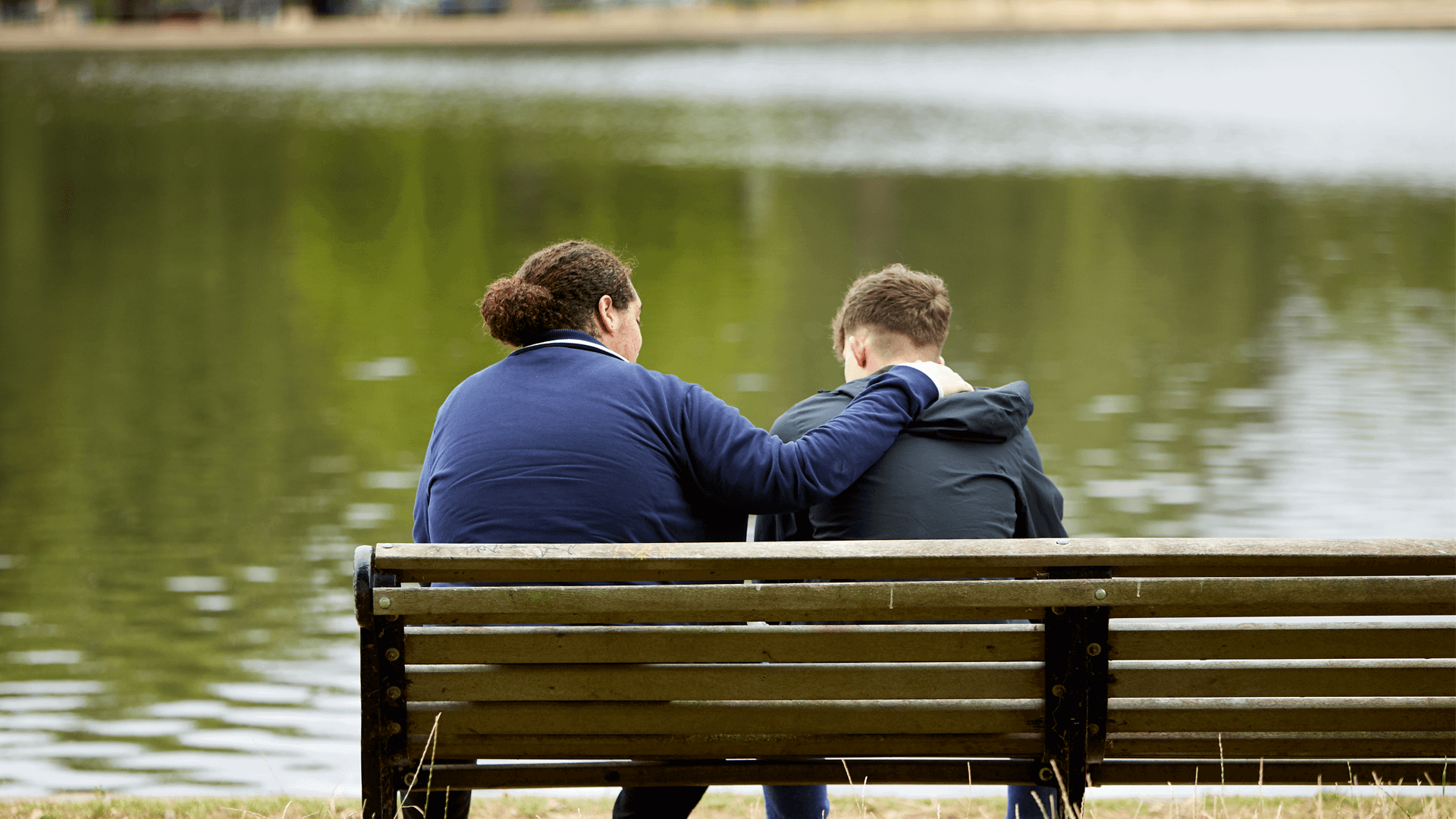 medium shot of two boys sitting on a bench in front of the lake comforting each other one boy puts his hand on the shoulder of his friend