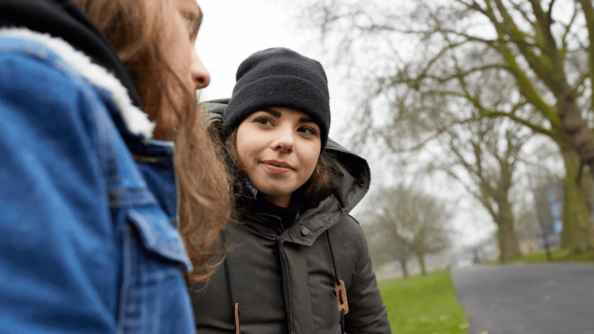 A girl in her winter coat is looking and smiling at her friend as they are talking while sitting on a park bench.