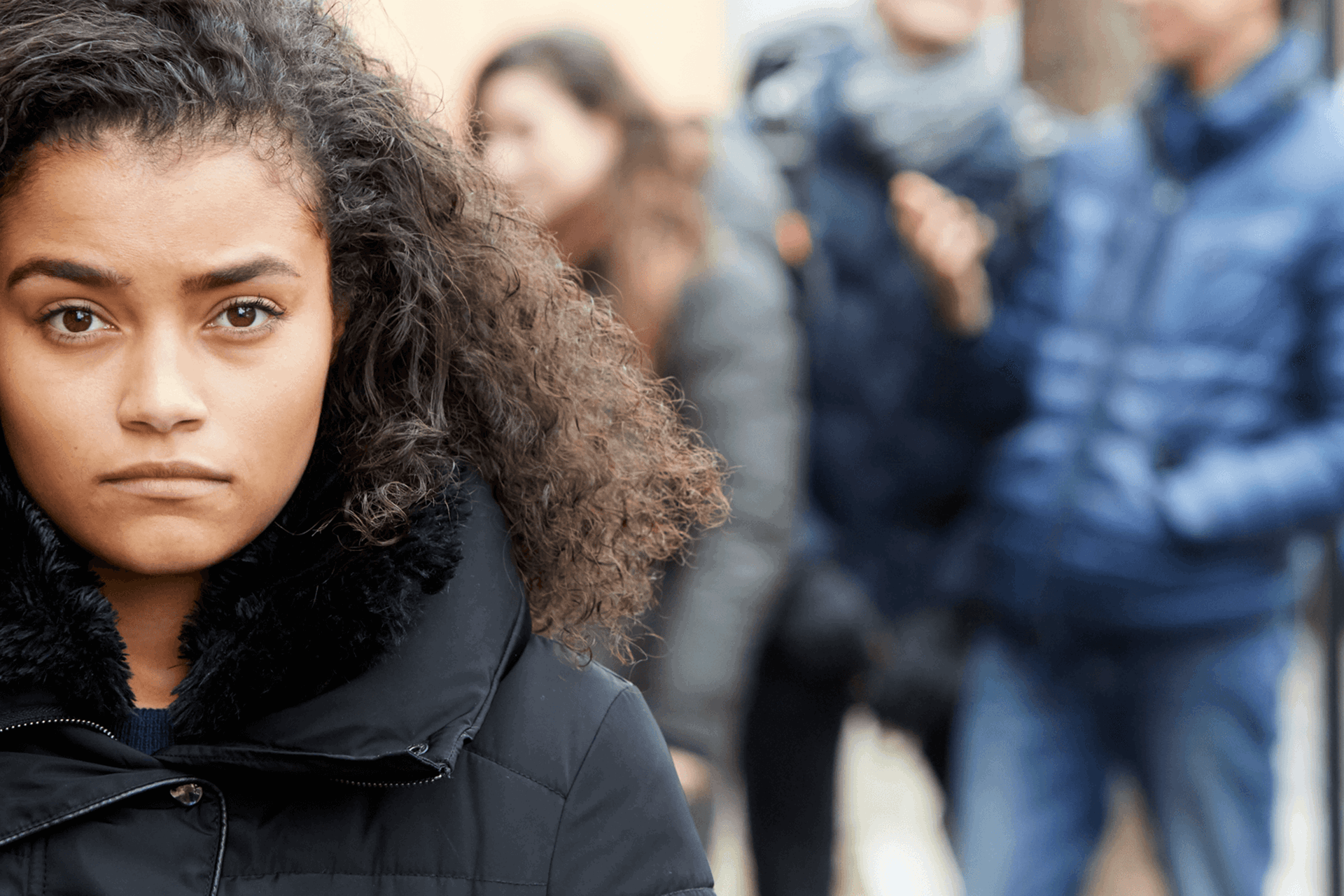 close up of a girl with a curly hair and wearing black jacket looking in front of the camera with group of young people on the background