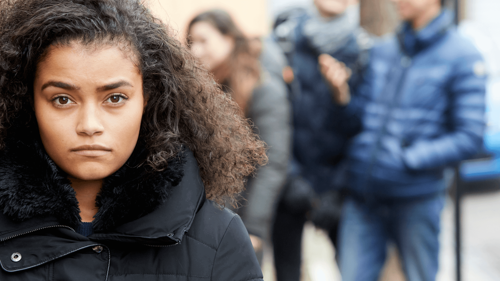 close up of a girl with a curly hair and wearing black jacket looking in front of the camera with group of young people on the background