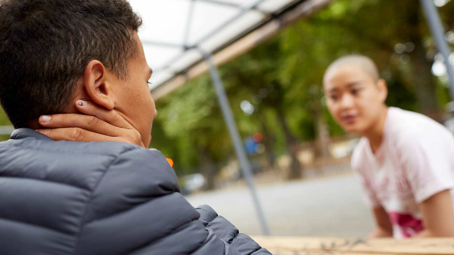 a-young-man-in-blue-jacket-with-face-unseen-talking-to-a-young-woman-with-shaved-head-in-white-shirt-while-sitting-on-a-bench-with-table-in-the-park