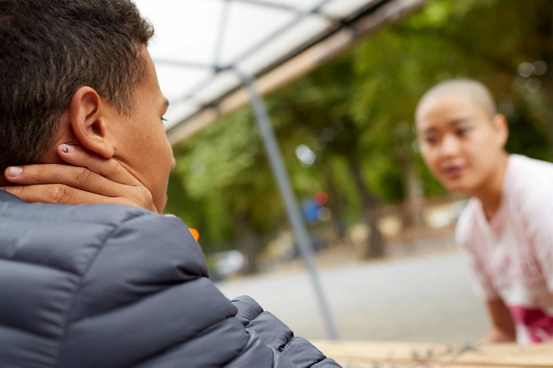 a-young-man-in-blue-jacket-with-face-unseen-talking-to-a-young-woman-with-shaved-head-in-white-shirt-while-sitting-on-a-bench-with-table-in-the-park