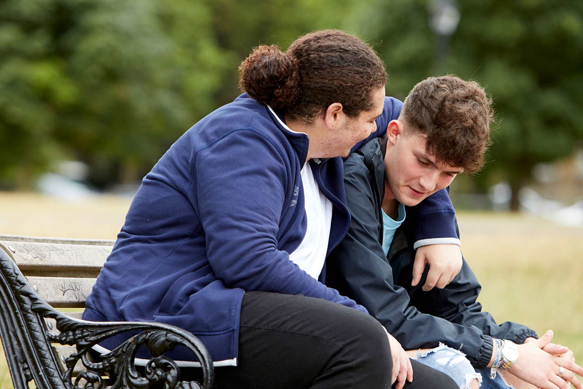 Two boys sit on a bench talking together in a park. They are smiling as one boy has his arm around the other's shoulder.