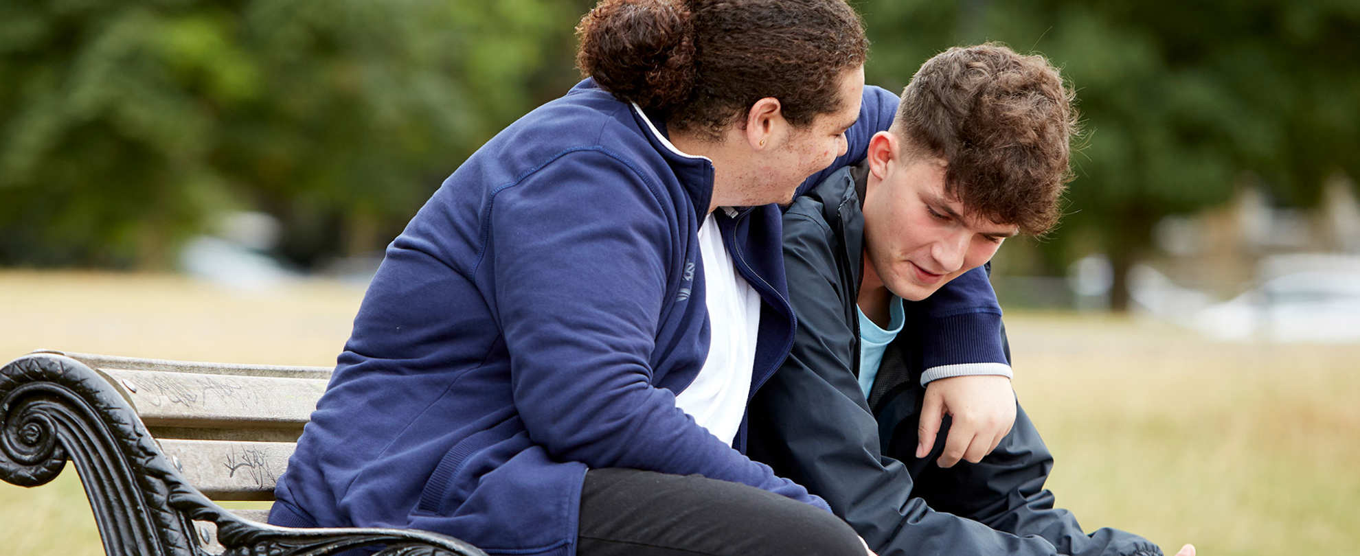 Two boys sit on a bench talking together in a park. They are smiling as one boy has his arm around the other's shoulder.