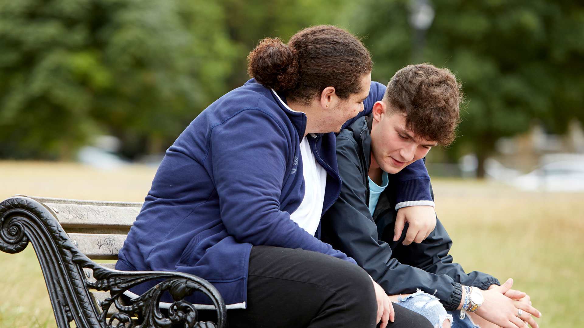 Two boys sit on a bench talking together in a park. They are smiling as one boy has his arm around the other's shoulder.