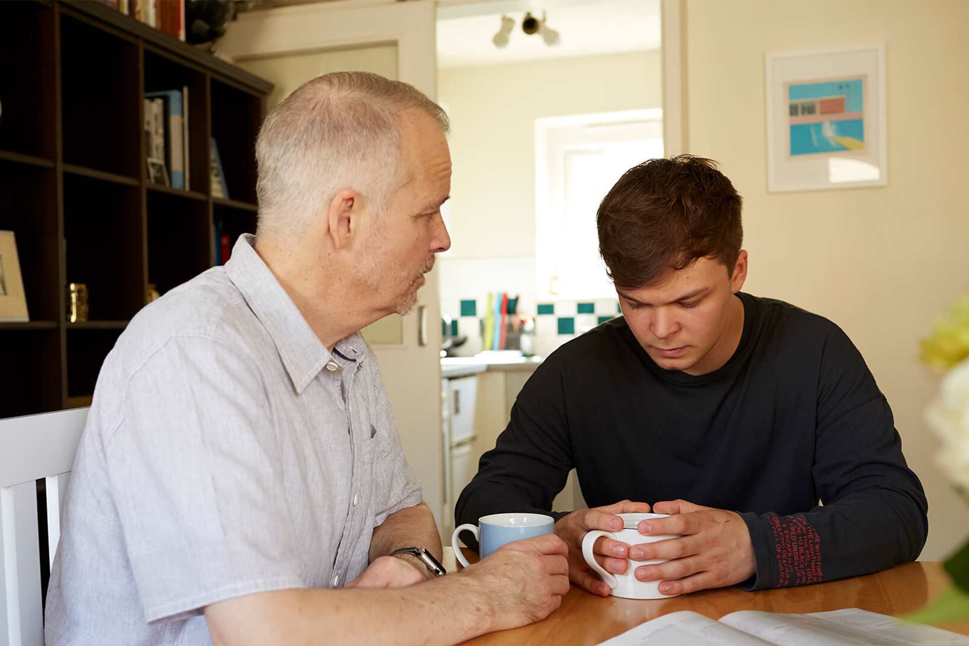 A father and son sitting at a table with hot drinks and serious facial expressions.