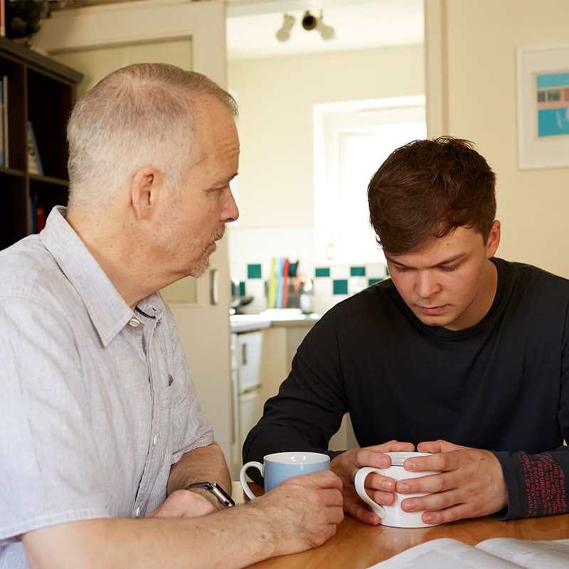 A father and son sitting at a table with hot drinks and serious facial expressions.