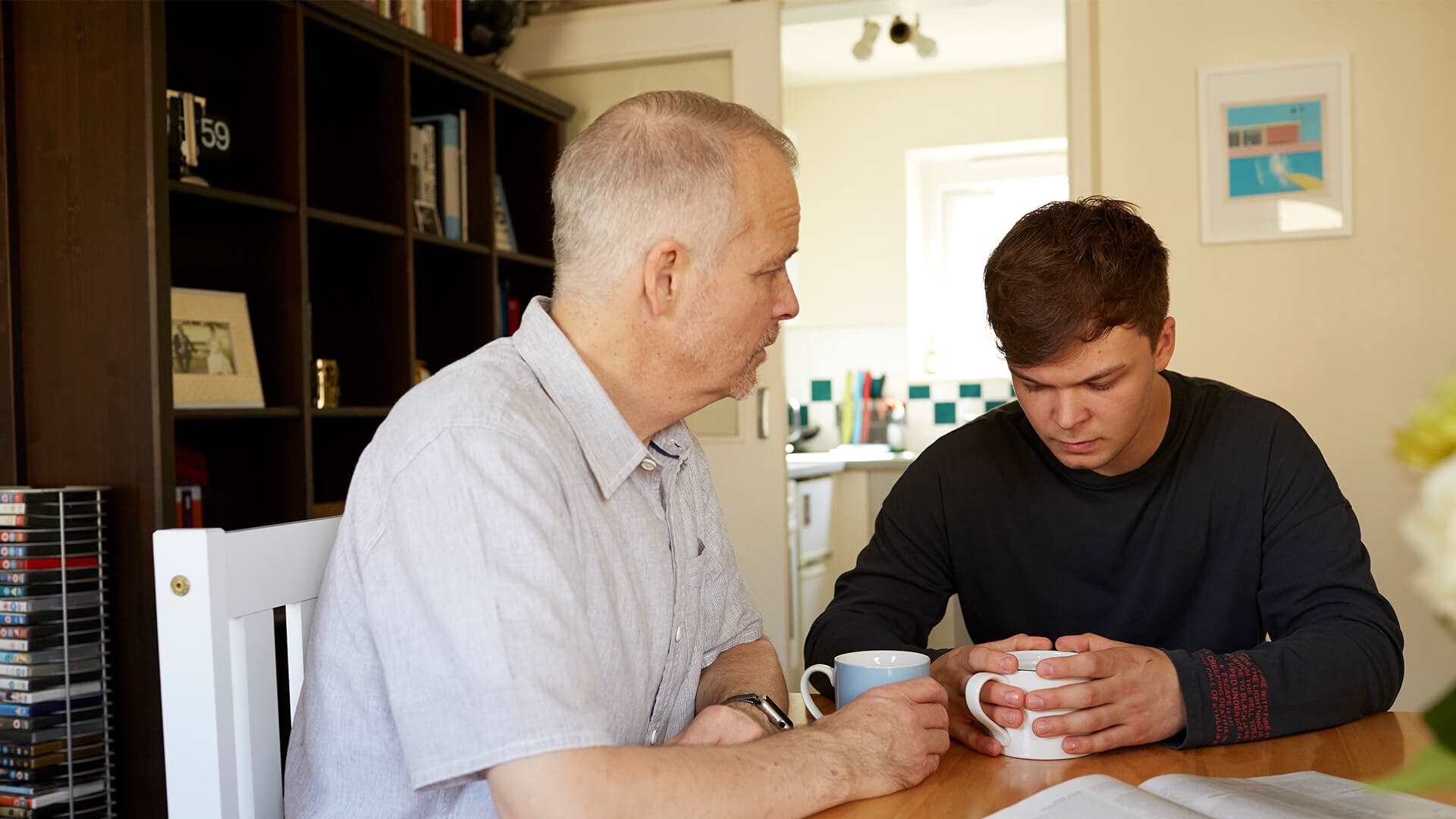 A father and son sitting at a table with hot drinks and serious facial expressions.