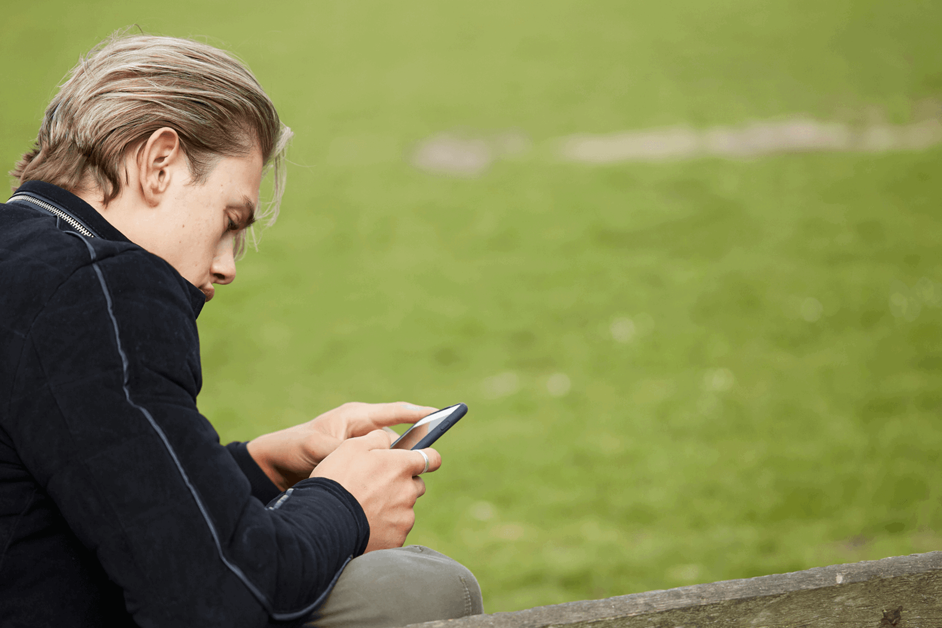 side view close up of a boy wearing black jacket looking at his phone while sitting on a bench in a park