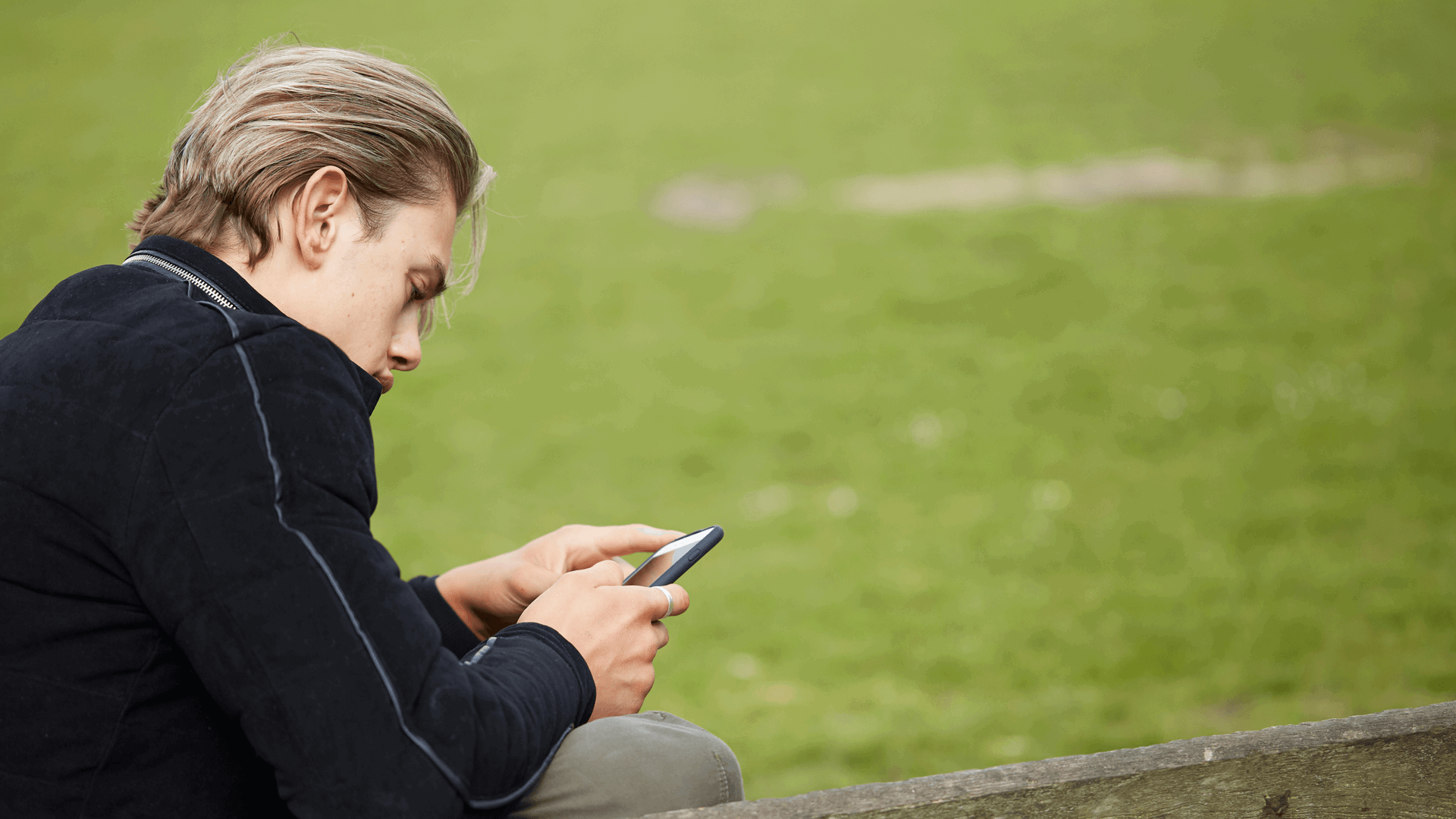 side view close up of a boy wearing black jacket looking at his phone while sitting on a bench in a park