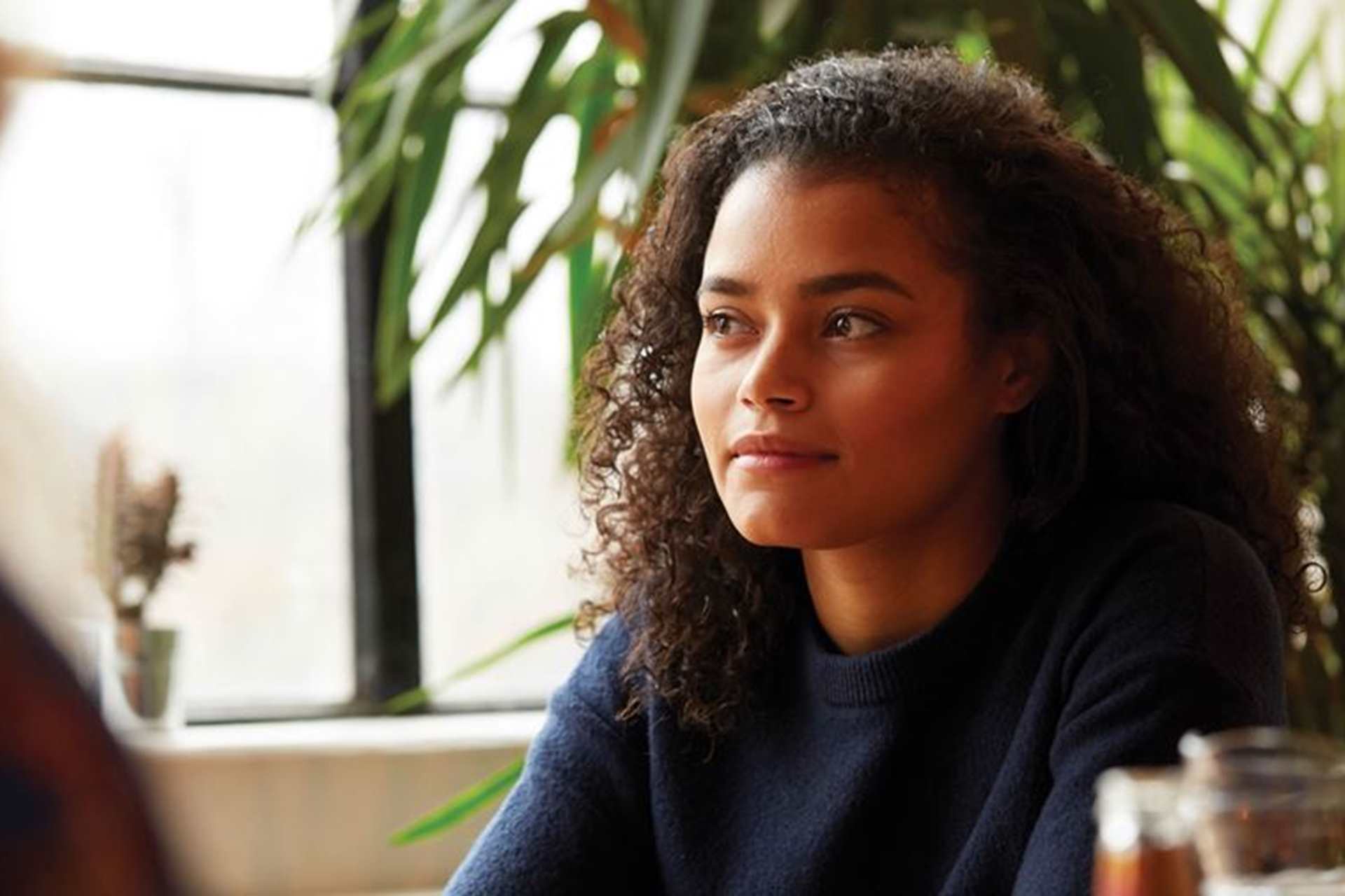 A girl with curly hair sitting in a cafe. She is looking away and smiling beside a window.