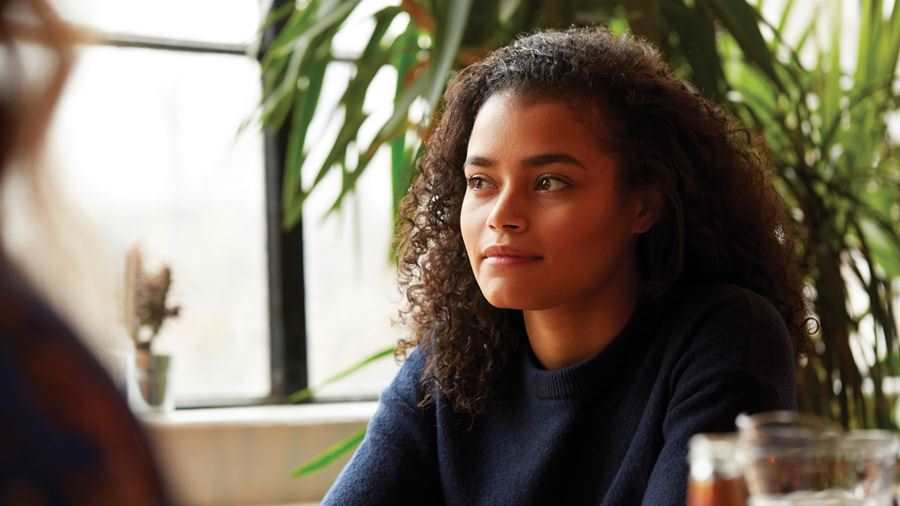 A girl with curly hair sitting in a cafe. She is looking away and smiling beside a window.