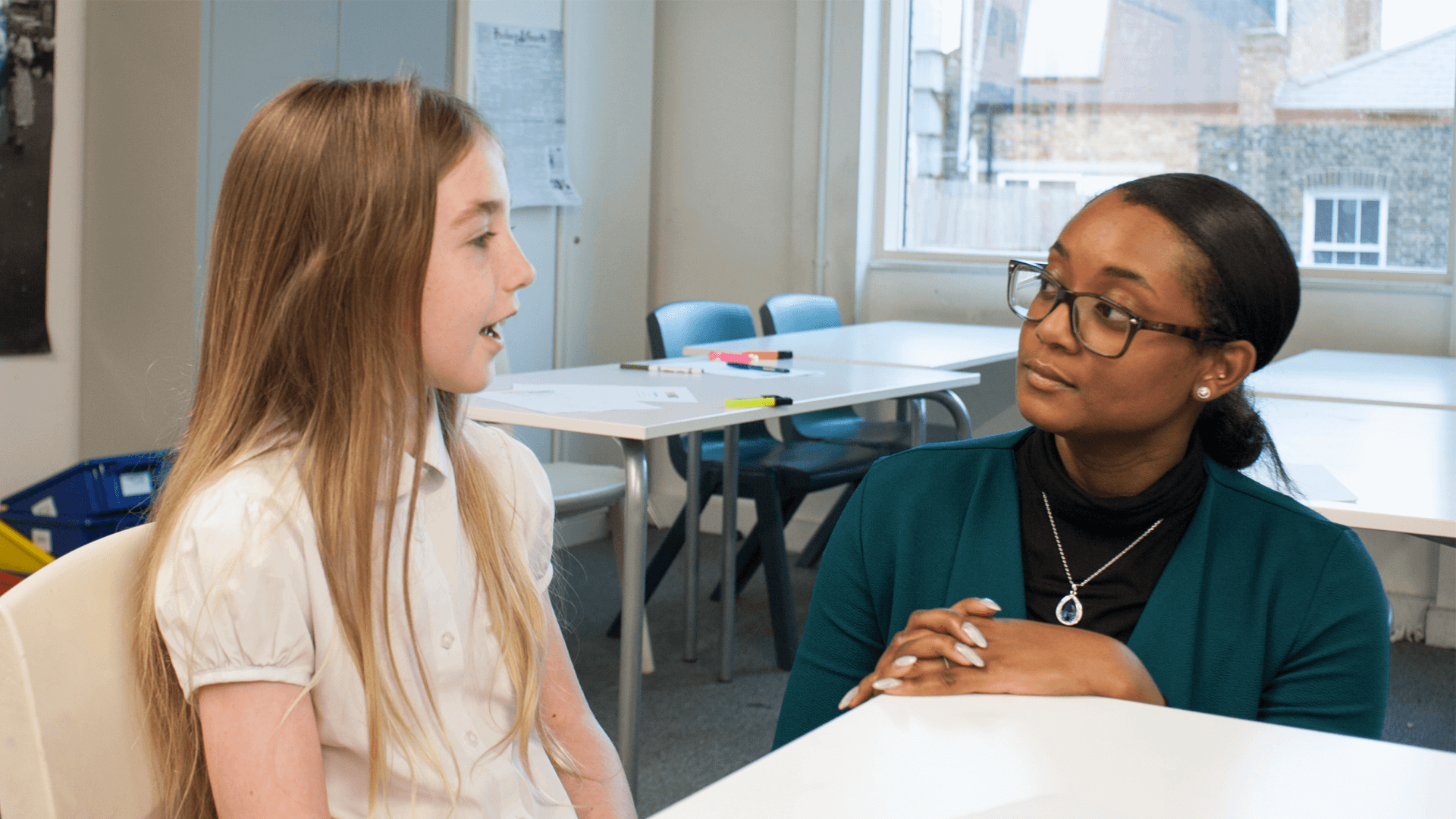 a girl student sitting on a school desk talking while a teacher sit beside her to listen