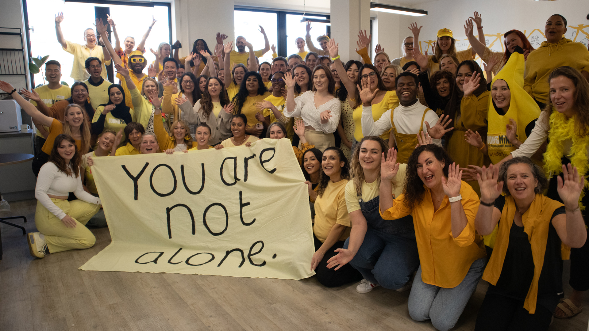 YoungMinds staff all wearing yellow for #HelloYellow 2023, posing with their hands in the air. They are holding a sign saying "You are not alone."