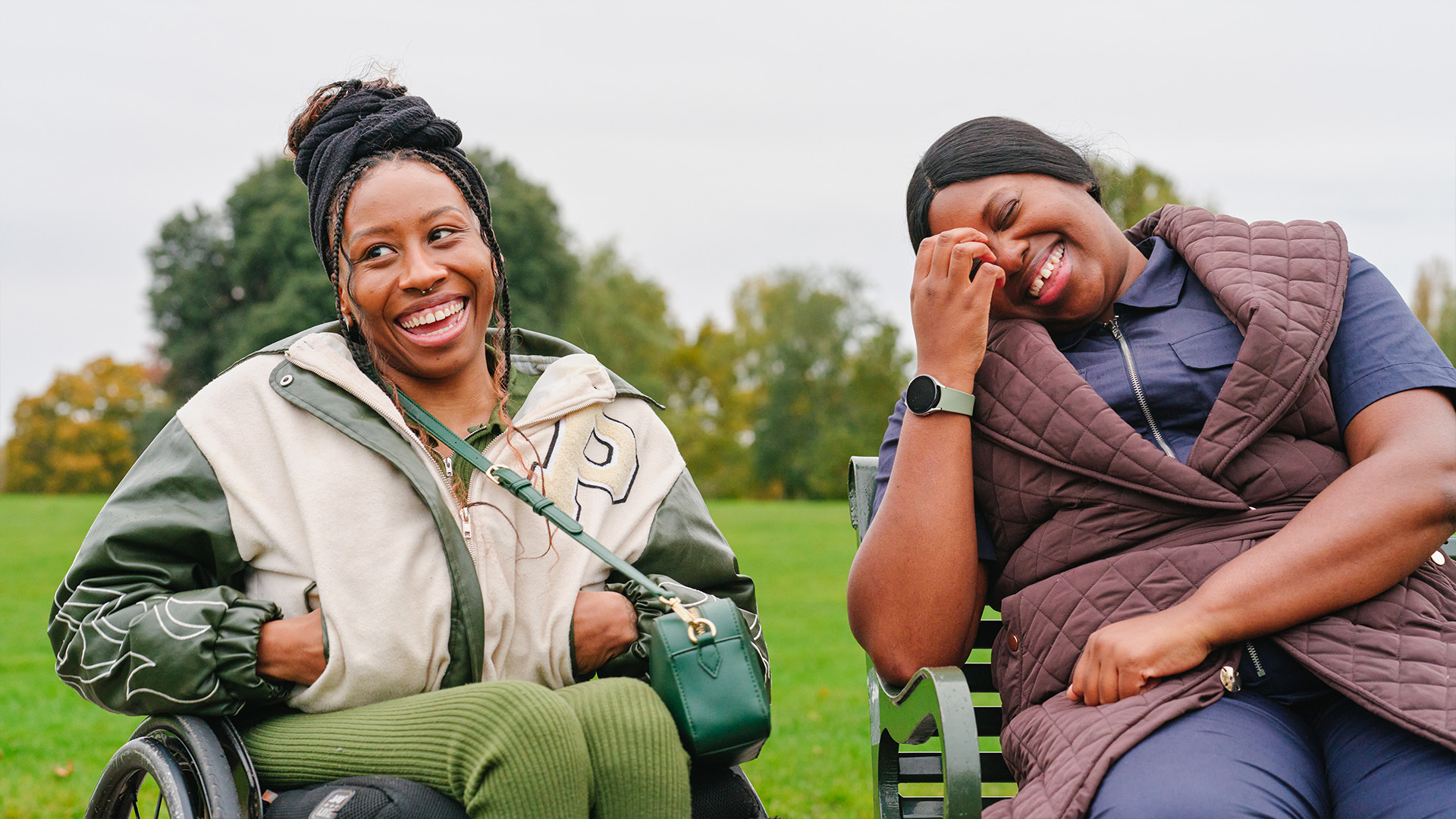 A young Black woman in a wheelchair and an older Black woman sitting on a bench in the park. They are laughing together.