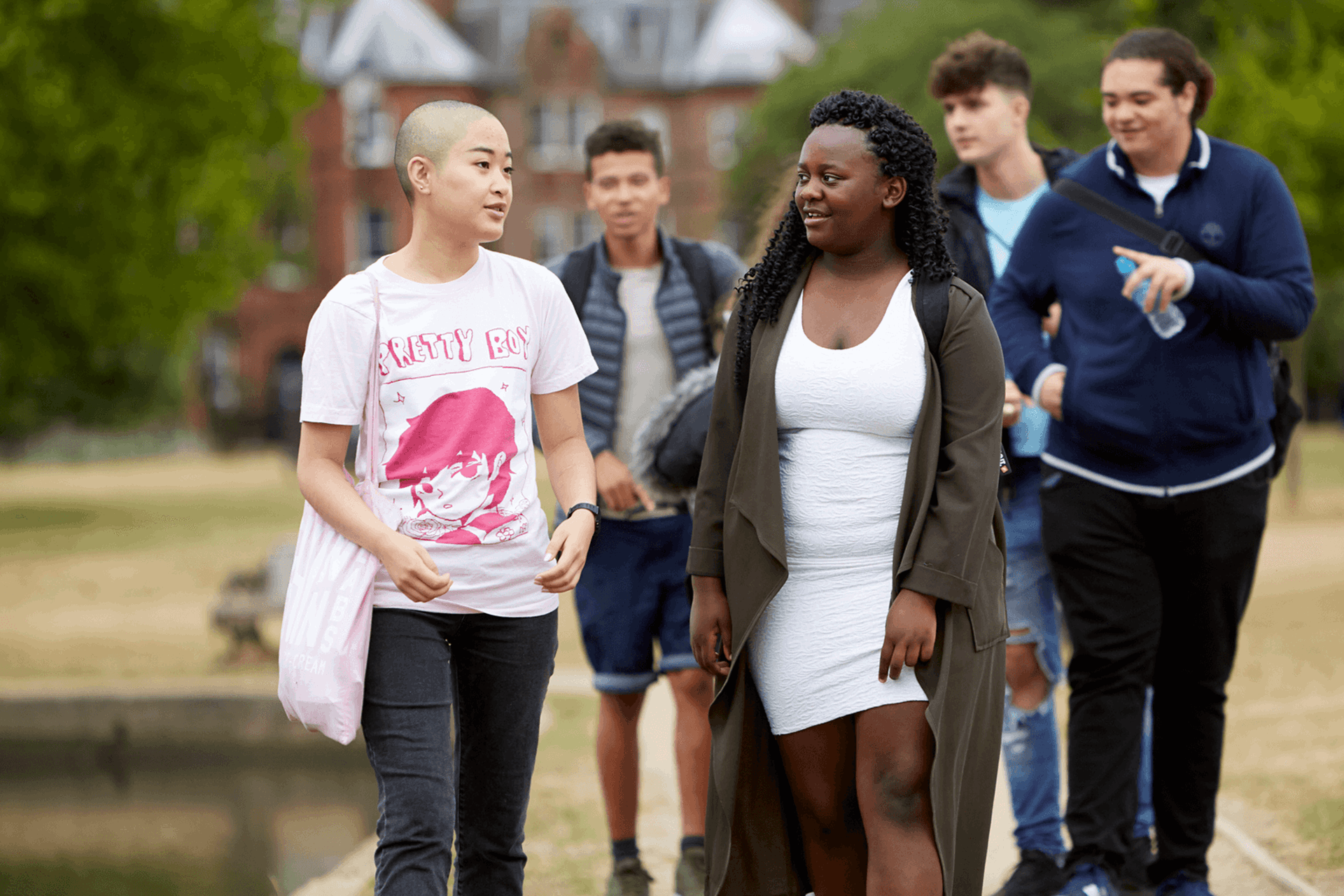 A group of five young people walking together along a path in the park. Two girls are walking ahead and talking, while three boys walk behind them.