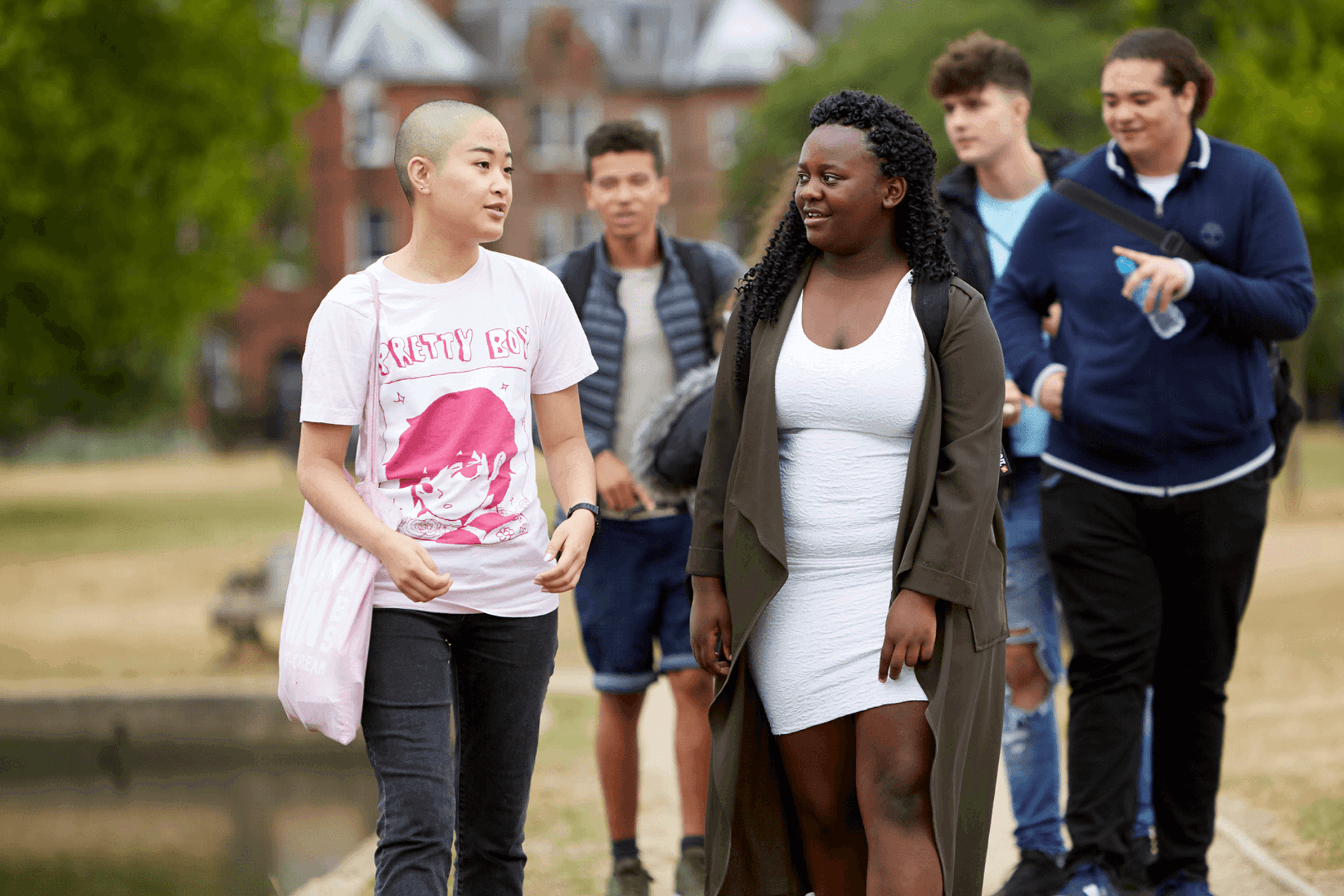 A group of five young people walking together along a path in the park. Two girls are walking ahead and talking, while three boys walk behind them.