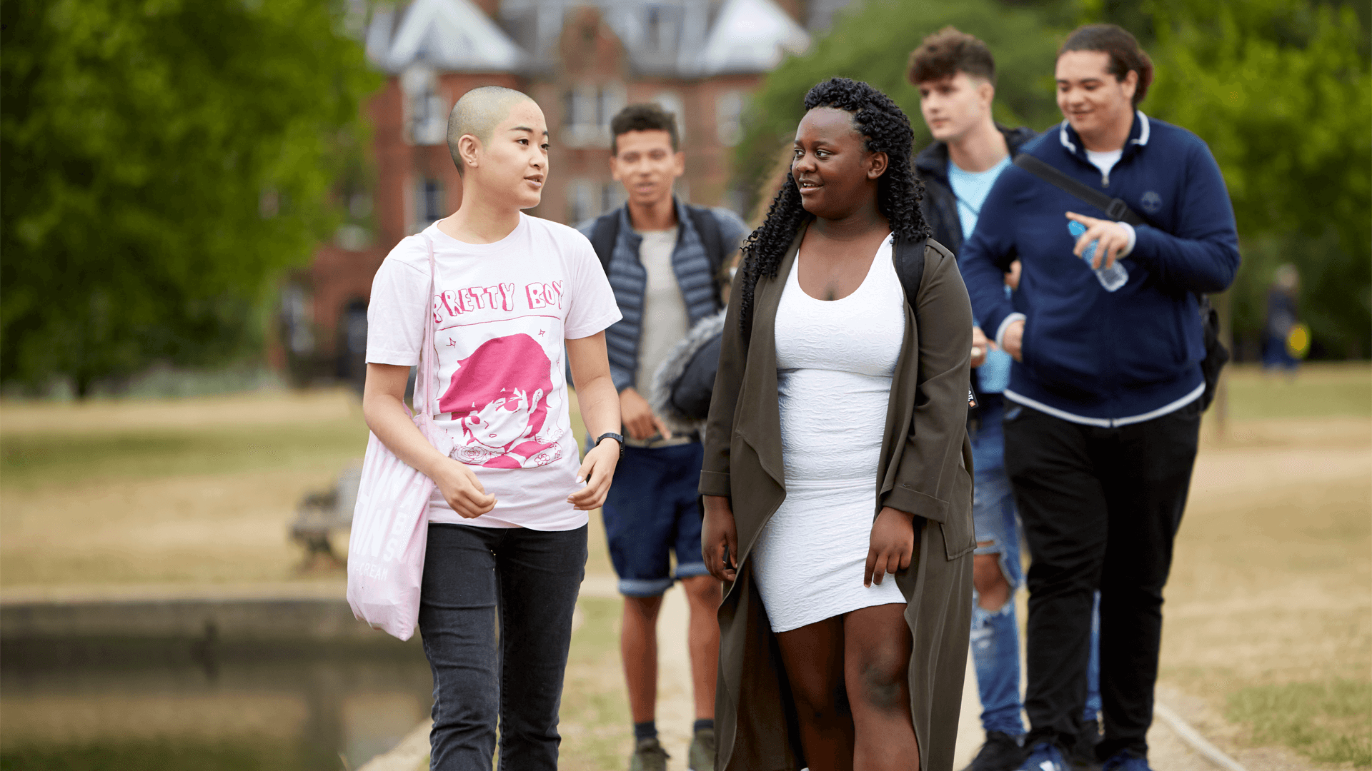 A group of five young people walking together along a path in the park. Two girls are walking ahead and talking, while three boys walk behind them.