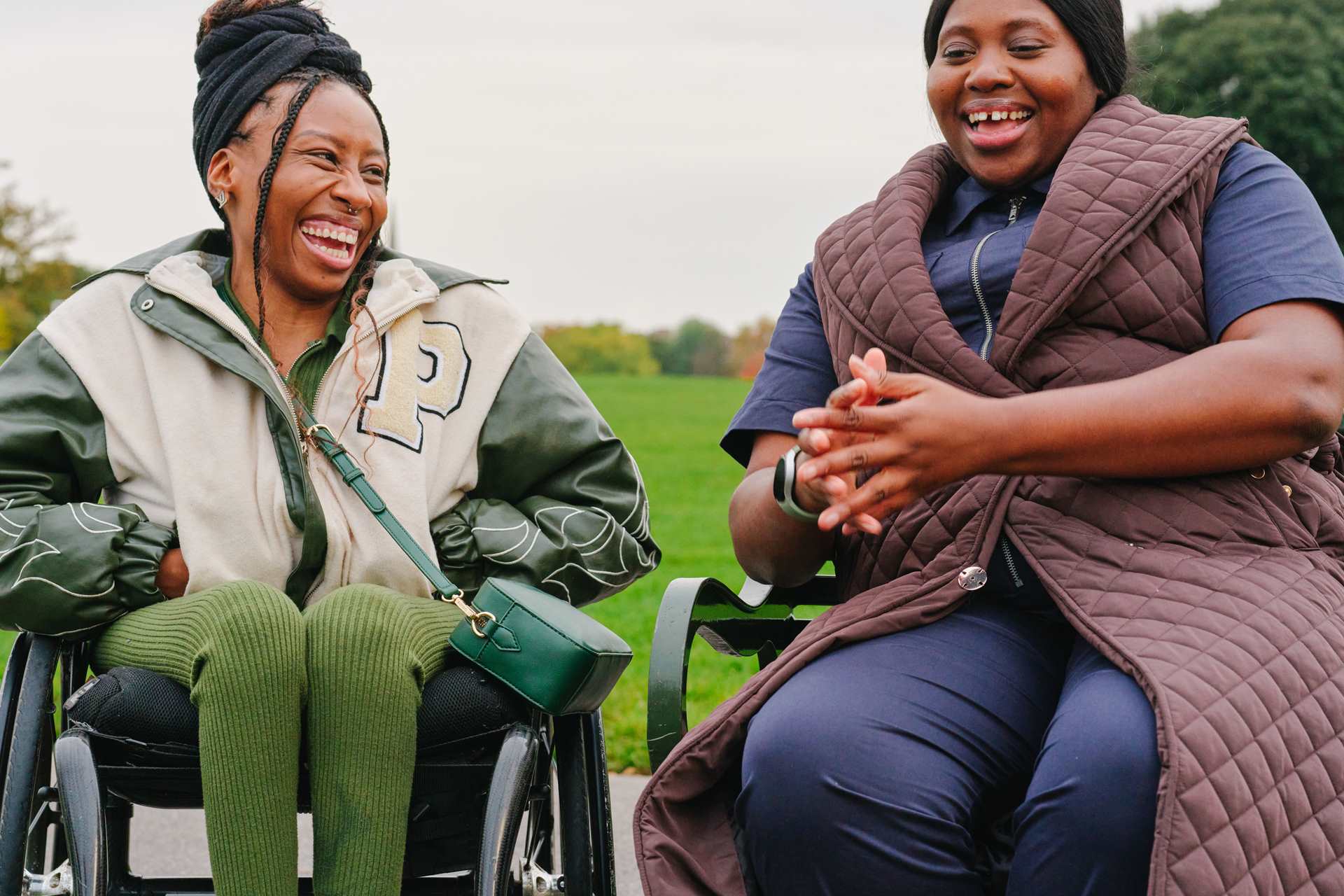 A young Black woman in a wheelchair and an older Black woman sitting on a bench in the park. They are laughing together.