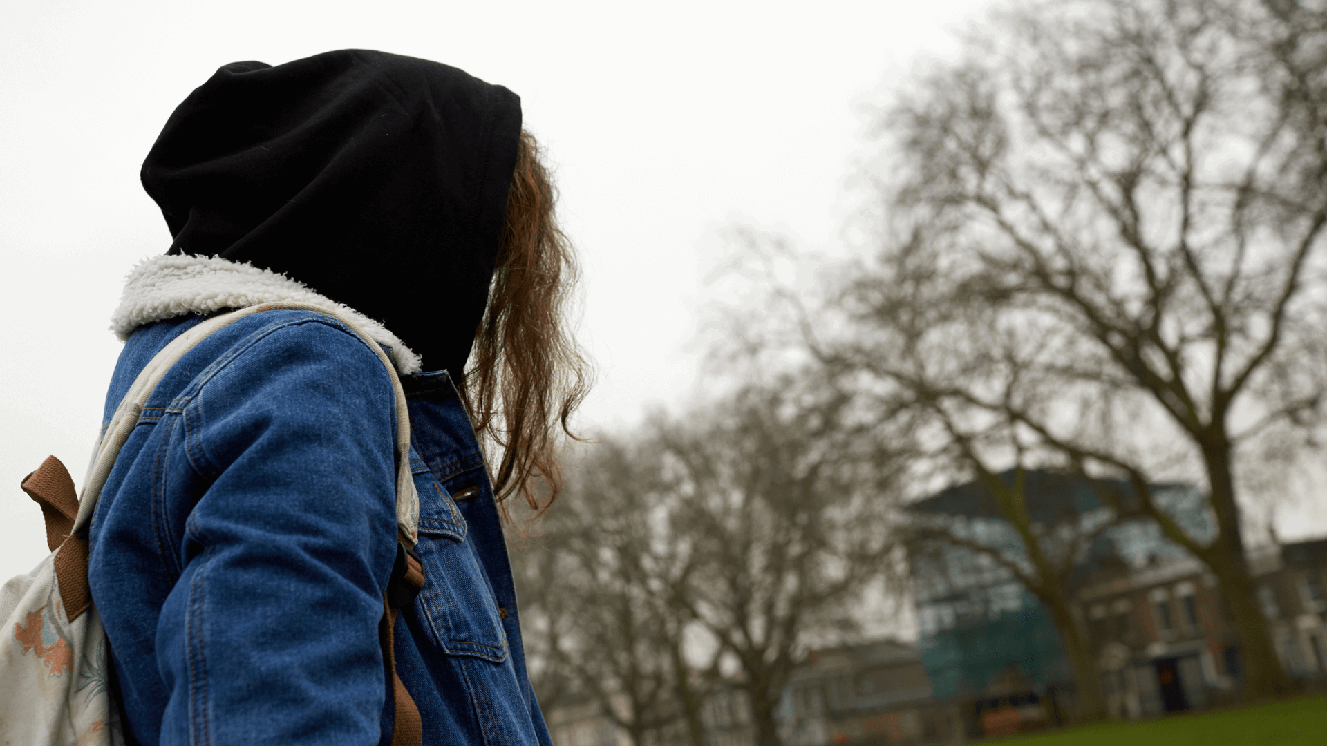 medium shot of a girl wearing a backpack and denim jacket hoodie and with face unseen while standing in park on a chilly weather 
